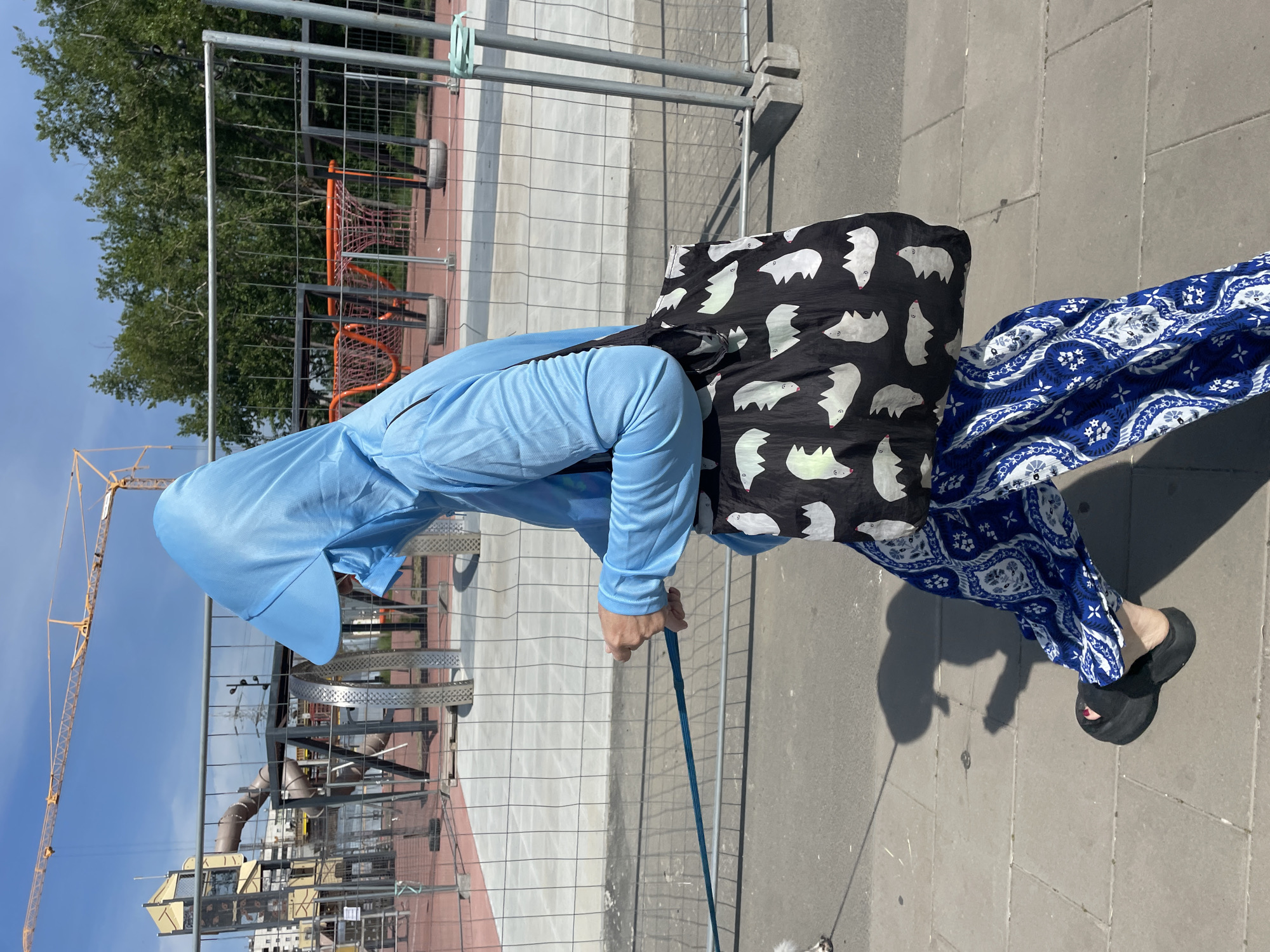 A person in a blue hooded jacket and blue patterned pants walks on a sidewalk, holding a leash attached to an unseen dog. The person carries a black tote bag with a white bat pattern. In the background, a construction site with a crane and playground equipment is visible behind a chain-link fence. The scene is illuminated by daylight under a clear blue sky.