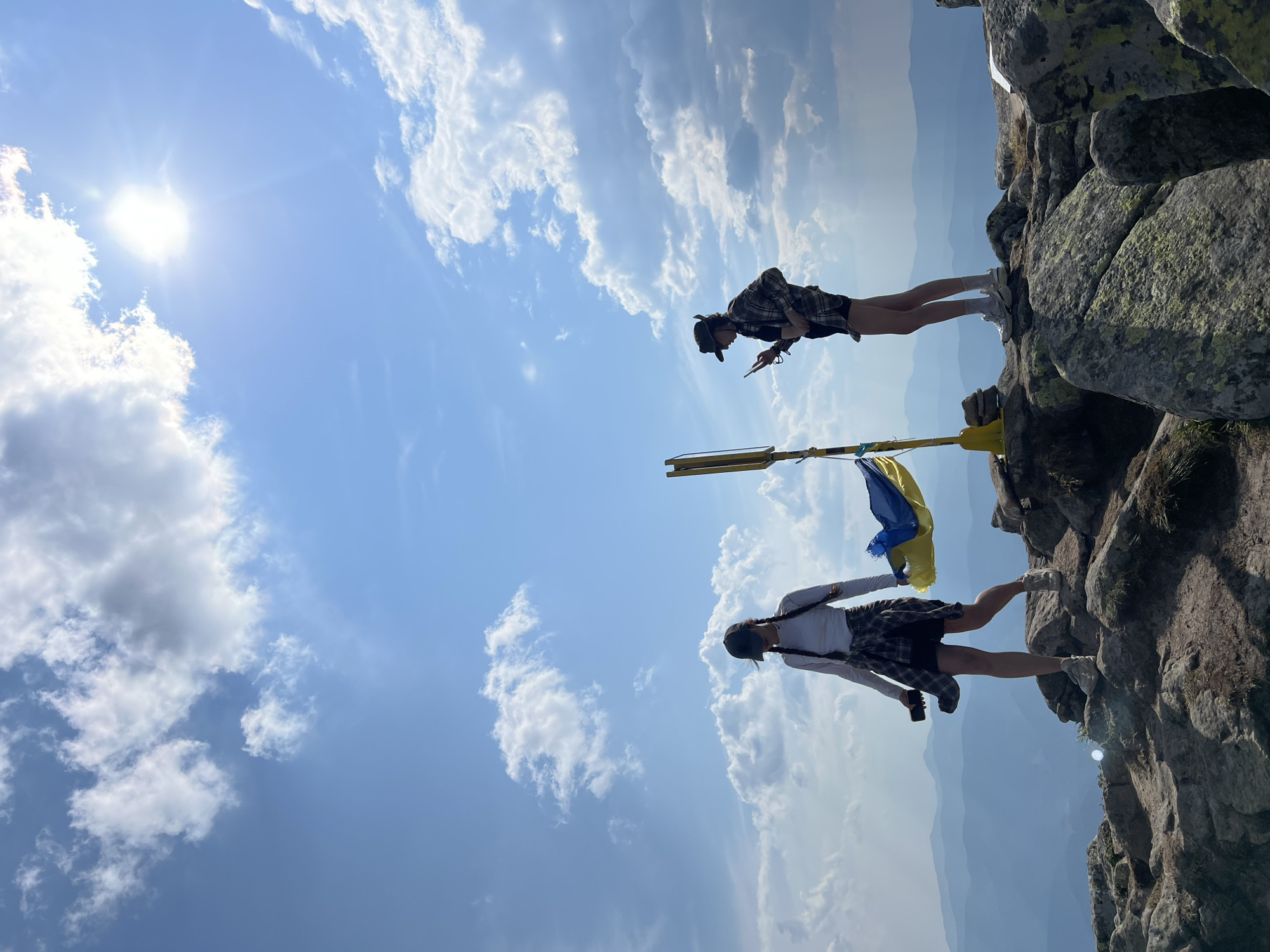 Two individuals stand on a rocky mountain summit, with a Ukrainian flag prominently displayed on a yellow pole. The person on the left, wearing a white long-sleeved shirt, a plaid shirt tied around their waist, dark shorts, a black baseball cap, and white sneakers, holds a smartphone in their right hand. Their left hand is extended, holding a dark object, possibly another phone. The person on the right, wearing a checkered shirt, dark shorts, a hat, and sneakers, is looking at their smartphone. The background features a vast expanse of layered mountain ranges fading into a hazy blue horizon under a bright, sunlit sky filled with dramatic clouds. The foreground consists of rugged, gray boulders and sparse vegetation. The scene appears to be the summit of Mount Bitkov, located in the village of Luhy, Zakarpattia Oblast, Ukraine.