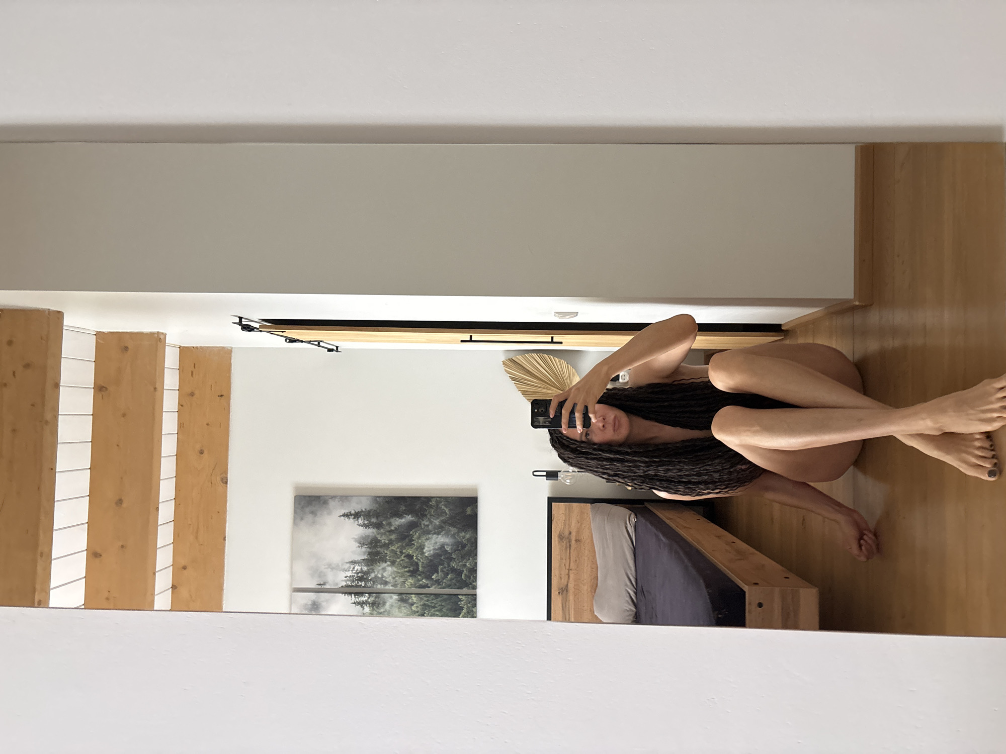 The scene, located in the city of Chaikovsky, Russia, depicts an adult female reflected in a full-length mirror, taking a selfie. She has long, dark dreadlocks and is seated directly on a light-colored wooden floor with her legs crossed at the ankles. She holds a black smartphone with both hands, obscuring her face from the nose up; her eyes are visible, showing a neutral expression. The woman appears to be nude, or wearing minimal dark undergarments, revealing her shoulders, chest, abdomen, and legs. Her toenails are painted with dark polish.
The background, visible through the mirror's reflection, shows an interior room with predominantly white walls and a ceiling featuring exposed light-colored wooden beams. On the wall behind the subject, a large, rectangular, vertically oriented two-panel artwork depicts a dense forest of dark green coniferous trees shrouded in mist or fog. To the left of the reflected subject, a simple wooden bed frame with a dark grey or navy blue fitted sheet and a light grey pillow is partially visible. A black lamp with an exposed bulb is positioned to the right of the bed. Above and to the right of the reflected subject's head, a golden, fan-shaped decorative object is mounted on the wall. A dark vertical element, consistent with a door frame or sliding barn door, is visible to the far right within the reflection, adjacent to a section of a light grey wall.