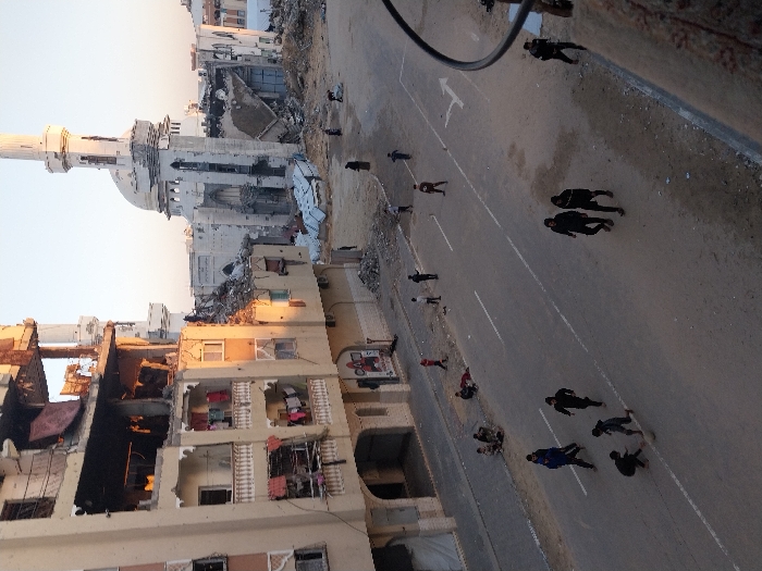 A severely damaged urban landscape shows buildings partially collapsed and riddled with holes, indicative of heavy conflict. A mosque's minaret stands relatively intact amidst the destruction. People walk along a street marked with directional arrows. The scene is desolate, highlighting the aftermath of violence, with scattered debris and damaged structures dominating the view. The angle suggests the image was captured from an elevated position.