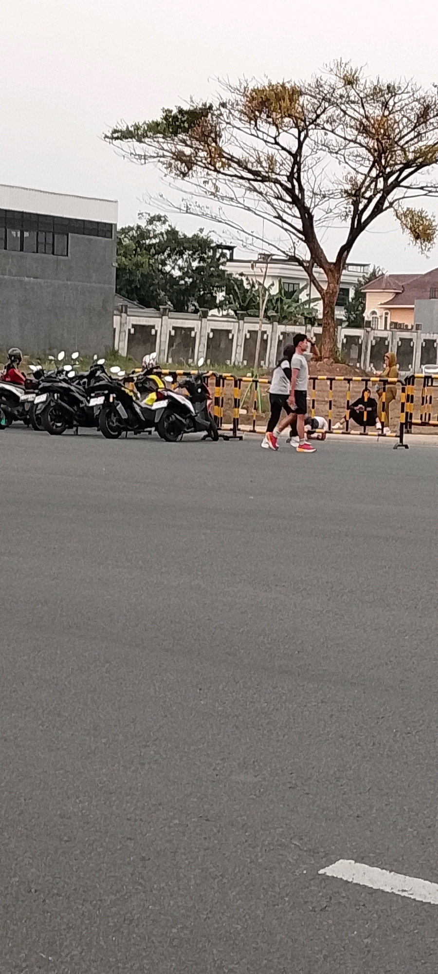 In Gunung Putri, Indonesia, a scene unfolds in front of a modern building and a row of parked motorcycles. Two individuals, a man and a woman, are walking side-by-side on a paved surface, with the woman's arm draped over the man's shoulders. The man is wearing a gray t-shirt and black shorts, while the woman is in a black and white striped shirt and black leggings. Both are wearing athletic shoes, the man's being red and white, and the woman's appearing to be white.
Behind them, a group of motorcycles are parked in a line, with several individuals sitting or standing near them. A man in a brown hooded jacket is visible on the right side of the image, and two people are seated on the ground behind a yellow and black barricade. The background features a building with dark windows and a large tree with sparse, autumn-colored leaves. The sky is overcast. The overall impression is a snapshot of everyday life in a developed urban or suburban area.