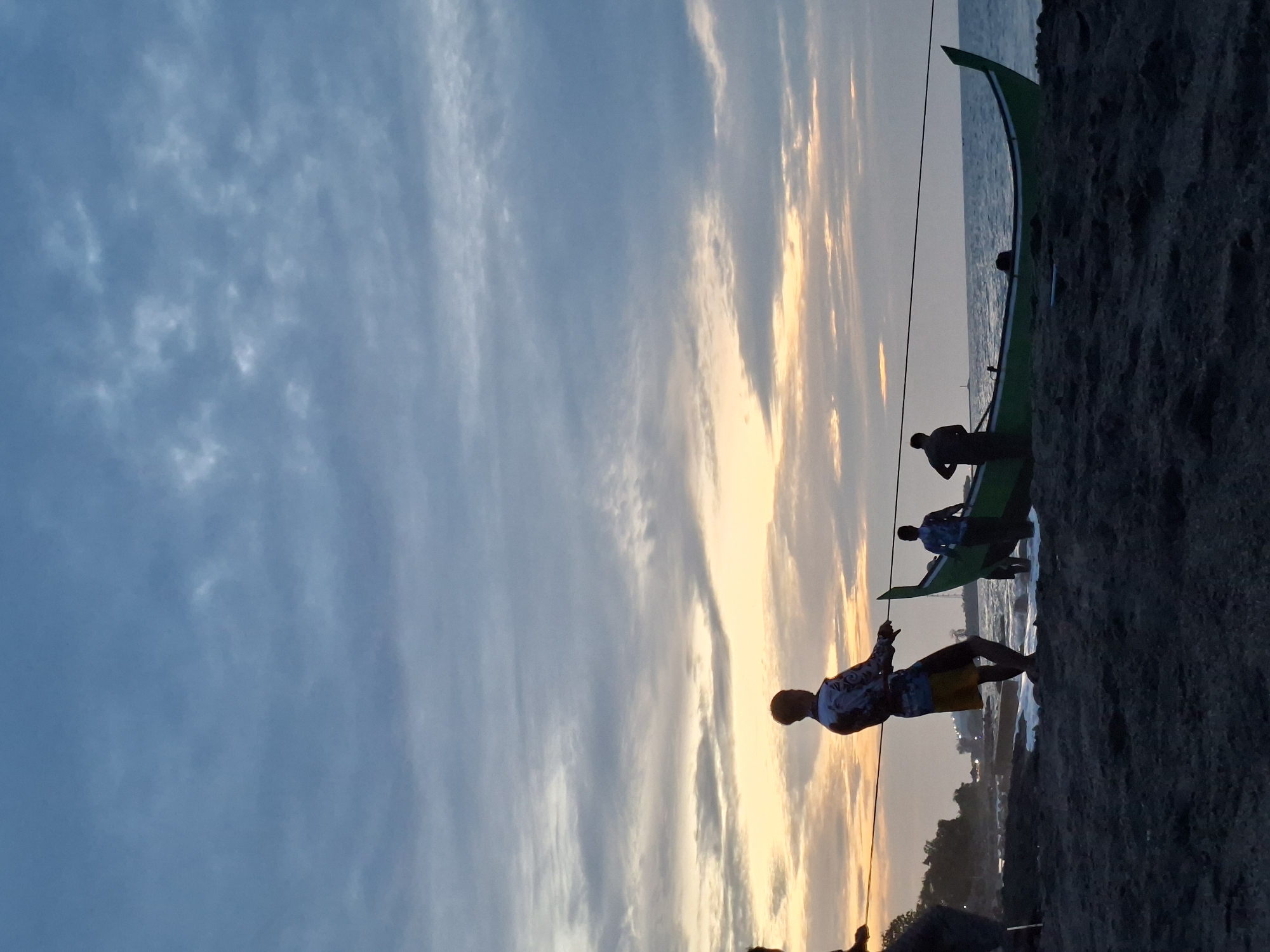 The image depicts a coastal scene at dusk, likely in Lhokseumawe, Indonesia, given the presence of traditional fishing boats and local attire. The sky is a dramatic blend of deep blues and muted oranges, with scattered clouds catching the last rays of sunlight. On a dark, sandy beach, several figures are silhouetted against the horizon. The primary focus is on a group of men pulling a green fishing boat ashore with ropes. One man in the foreground, wearing a patterned shirt and yellow shorts, is actively pulling a rope, his body angled with exertion. Behind him, other men are also engaged in the effort, some inside the boat and others on the shore. The water laps gently at the edge of the beach, and in the distance, a coastline with scattered buildings and vegetation can be seen. The overall atmosphere is one of community effort and the close of a day's work.