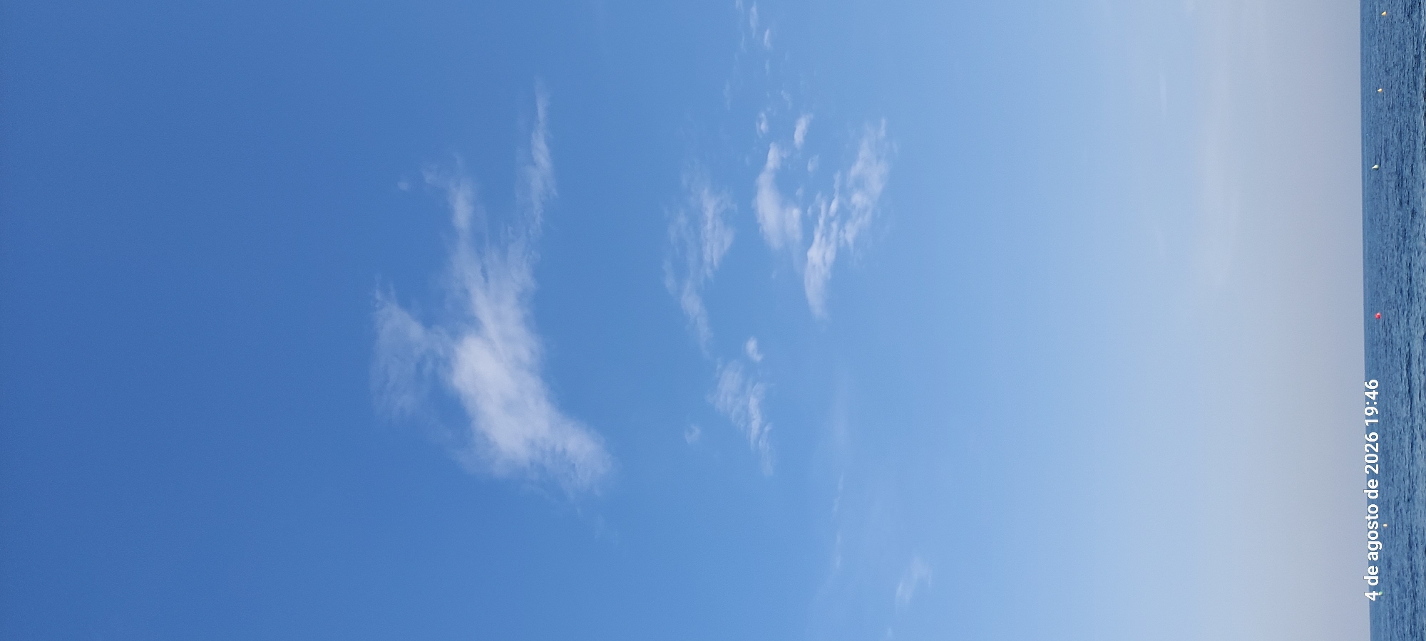 A wide shot captures the vast expanse of a bright blue sky with sparse, wispy white clouds. The horizon line shows the dark blue sea with a few small buoys. The lower left corner displays a timestamp: "4 de agosto de 2026 19:46", indicating the image was taken on August 4, 2026, at 7:46 PM. The scene depicts a calm sea and sky, consistent with an evening view from the coast of Castelldefels, Spain. There are no people or prominent structures visible in the frame.