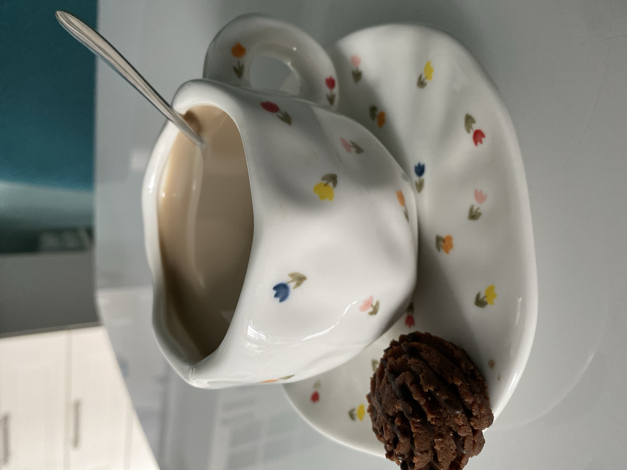 A close-up studio shot features a floral-patterned tea cup and saucer. A small, dark chocolate cookie rests on the edge of the saucer. A metal spoon is placed inside the tea cup. The background suggests a modern interior with a turquoise accent wall and white cabinetry. The image is rotated to the right. The location is the city of Unknown, Unknown.
