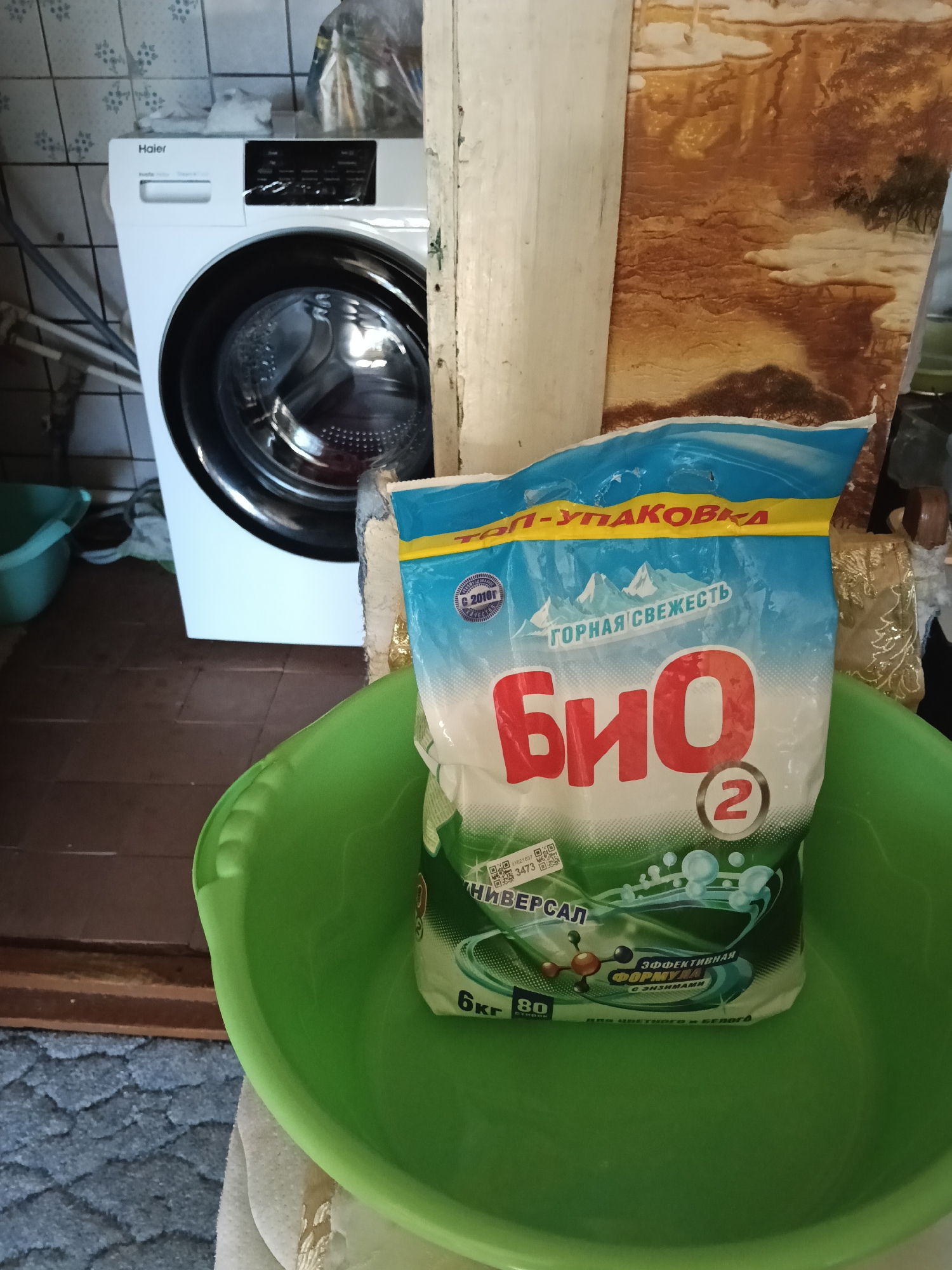 The image captures a domestic scene in Красный Маяк, Russia, presenting a Haier washing machine situated near an interior wall and a green basin containing a package of "BIO" washing powder. The washing machine is positioned to the left, and the green basin is situated in the foreground, holding the washing powder. The wall to the right appears to have a decorative pattern resembling a landscape. The floor is covered with tiles or laminate.