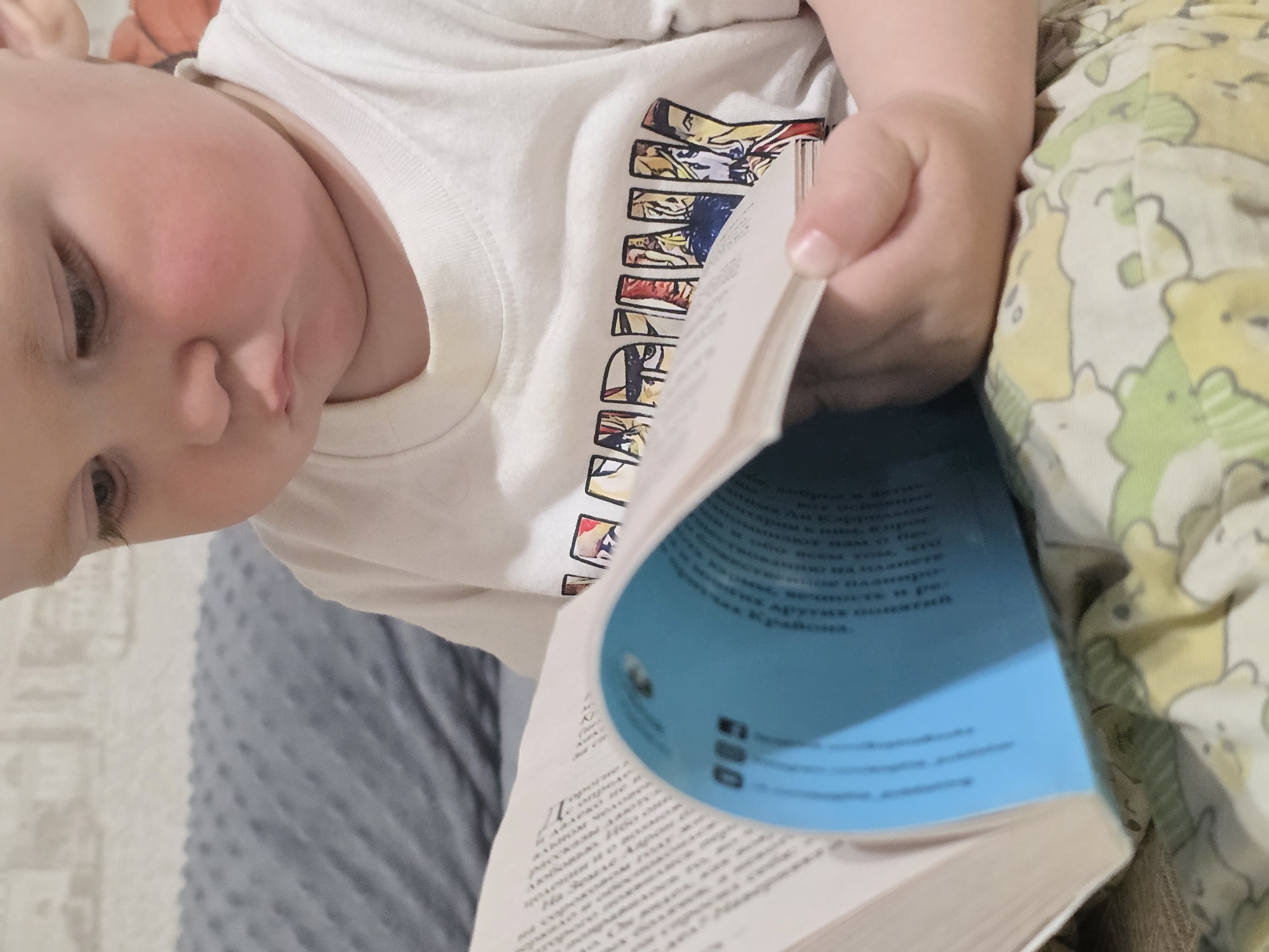 A baby is sitting on a gray and white patterned surface and a bed with a light yellow and green bear pattern, holding and seemingly reading a book. The book has a light blue cover, and its pages are open, revealing text in a language that cannot be determined from the image. The baby is wearing a white t-shirt with a colorful graphic print on it. The location and city are not discernible from the image, so the claim that it's in Chaikovsky, Russia, cannot be verified.