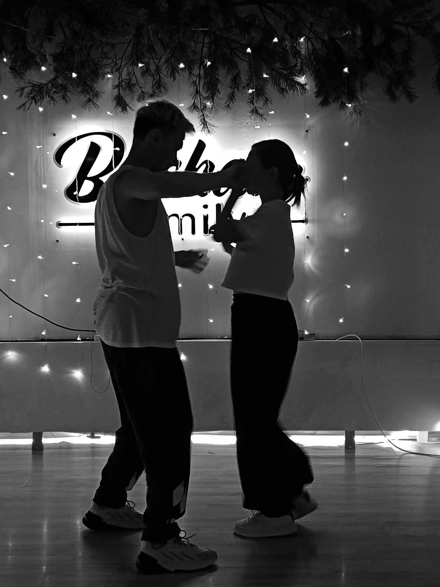 The image depicts a silhouetted couple in front of a backdrop with a neon sign reading "Bobo smile" and strings of lights. Greenery hangs above. The man is wearing a tank top and sweatpants, while the woman is wearing a T-shirt and similar pants. They appear to be interacting closely, possibly kissing or embracing. They are standing on a glossy floor. Based on the neon sign, the location may be related to the “Bobo Smile” brand and is possibly located in the city of Chaikovsky, Russia.