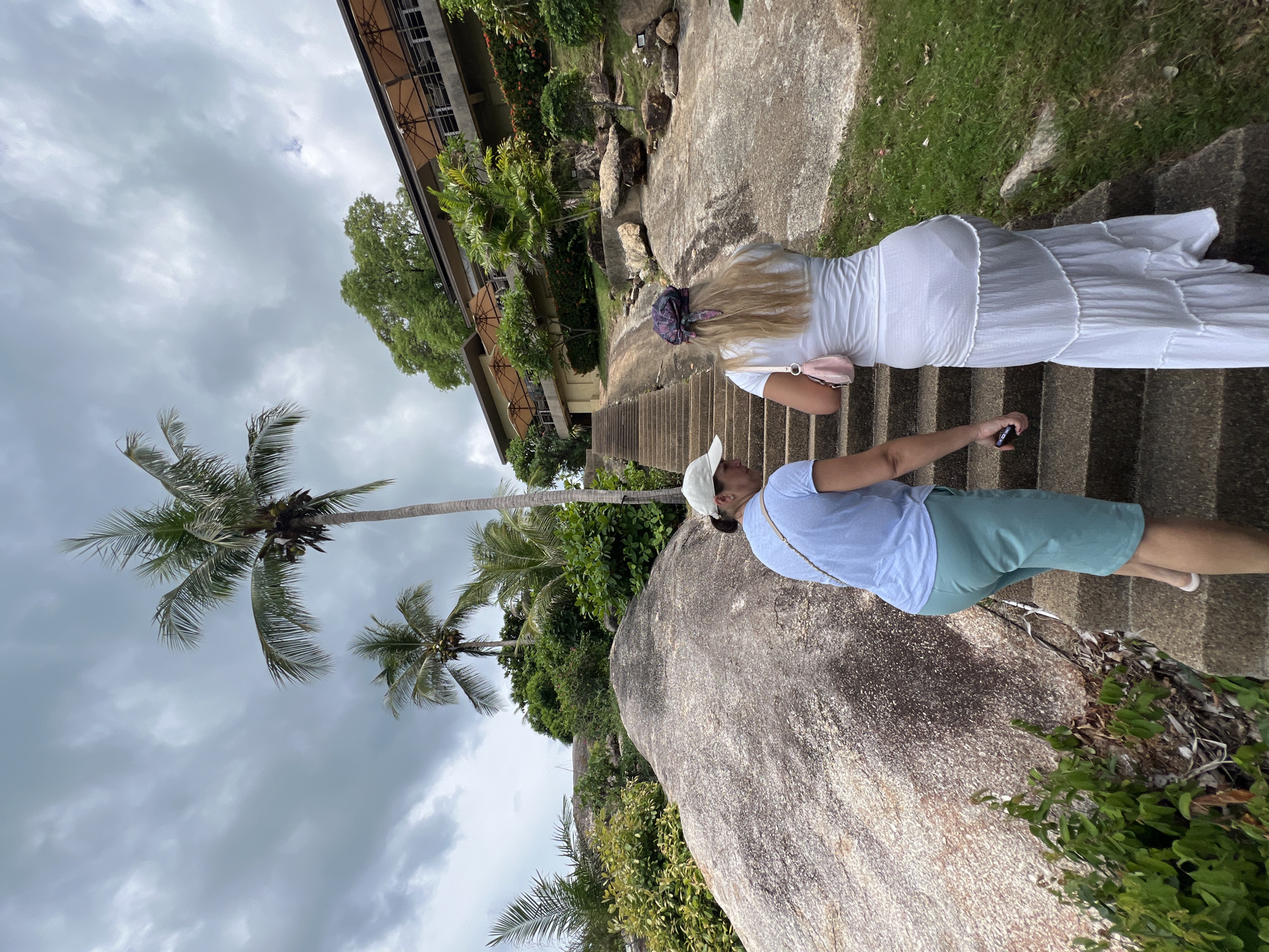 Two individuals are walking up a stone staircase in a tropical outdoor environment. The person in front is wearing a white dress and a headscarf. The person behind them is wearing a blue t-shirt and teal shorts, and is holding a phone. The staircase is surrounded by large boulders, lush green vegetation, and palm trees. In the background, a building with an outdoor seating area and umbrellas is visible, situated on a hillside. The sky is overcast with dark clouds. The scene appears to be located in Baan Chaweng Noi, Thailand.