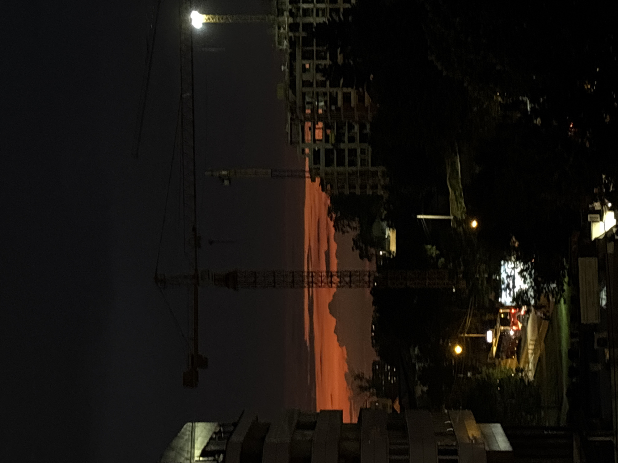 The image depicts a night scene in Belgrade, Serbia, featuring construction cranes against a dark sky with an orange glow, possibly from sunset. A building under construction is partially visible. Streetlights and building lights illuminate parts of the scene, including trees and nearby buildings.