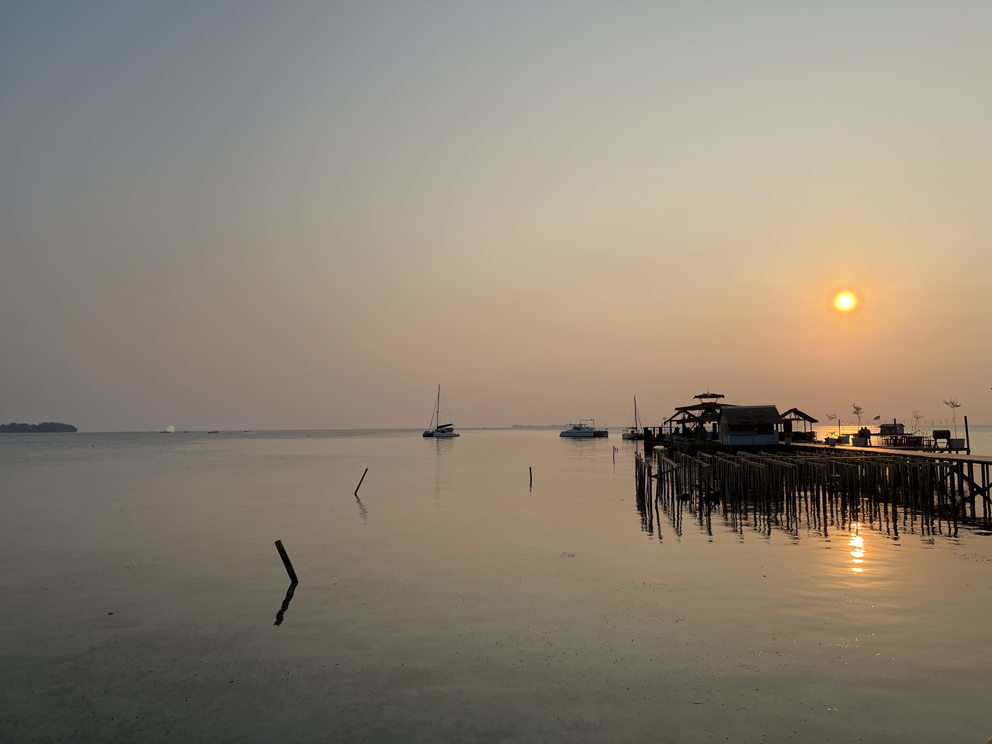 This photograph captures a serene sunset over the waters of Karimunjawa, Indonesia. The scene is dominated by the calm sea reflecting the warm hues of the setting sun. Several boats, including sailboats and smaller motorboats, are dotted across the water. A wooden pier extends into the sea, featuring structures that appear to be restaurants or accommodations. The pier is supported by numerous pilings, casting long shadows on the water's surface. The foreground shows the shallow seabed with a few submerged posts. The sky transitions from a pale blue at the top to a soft orange near the horizon, where the sun is a bright, luminous orb. The overall atmosphere is tranquil and picturesque, indicative of a peaceful coastal evening.