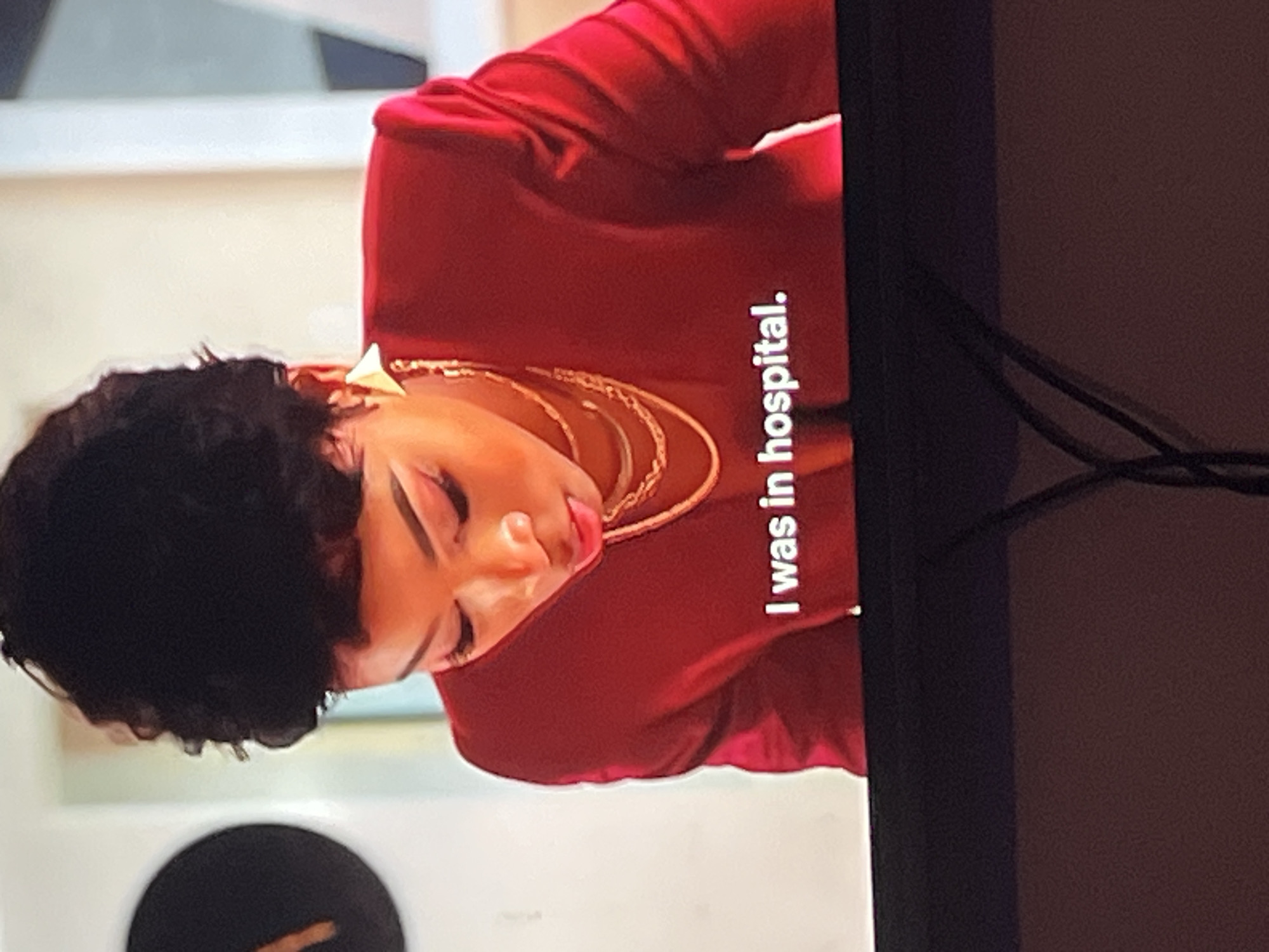 The media file shows a close-up shot of a woman wearing a red top and layered necklaces. She has dark, curly hair and is wearing makeup, including noticeable eyeshadow and lipstick. Her head is tilted down, and her eyes appear closed or looking downwards. The woman is looking towards the right side of the frame.
On the right side of the image, vertical white text is visible against the red background, reading "I was in hospital." The background behind the woman is out of focus, but appears to be an indoor setting with light-colored walls and some abstract shapes. The overall lighting suggests an indoor environment, possibly during the day or with artificial lighting. The woman's expression seems neutral or possibly conveys a sense of weariness or reflection. No specific time of day or weather conditions can be determined. The location context provided is Gura Top, Nigeria, but there are no specific visual cues within the image to confirm this location.