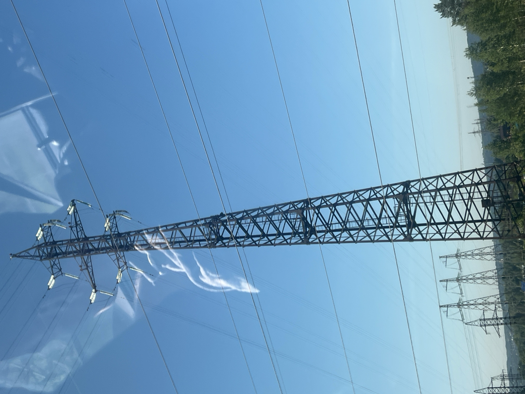 The image shows a tall, metal electricity pylon against a clear blue sky. Several power lines stretch horizontally across the frame. To the left of the main pylon, a distorted reflection is visible, possibly from a window. Wispy white cloud formations or contrails are present in the sky near the top of the pylon. In the background, a series of similar pylons are visible, receding into the distance, along with a treeline and a hilly landscape. The scene appears to be outdoors, under daylight conditions. The location is identified as Noviy, Russia.