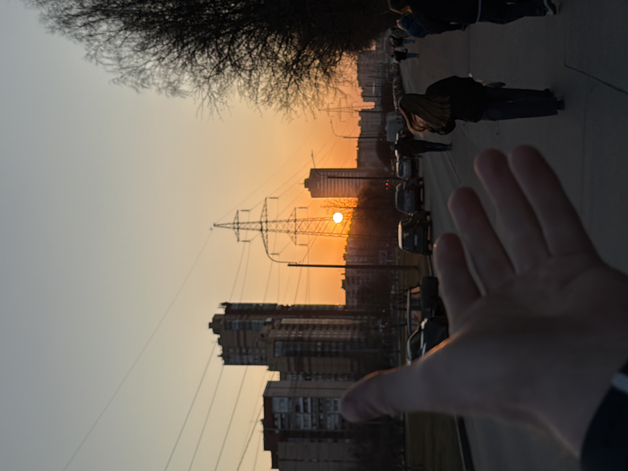 The image shows a sunset scene in what appears to be a city environment, possibly Saint Petersburg, Russia. The buildings suggest an urban setting, and the power lines could be part of the city's infrastructure. The presence of a hand in the foreground indicates the image was taken by a person, possibly reaching out towards the sunset. There are multiple cars parked along the side of a street or road, and several pedestrians can be seen walking, suggesting an everyday urban environment.