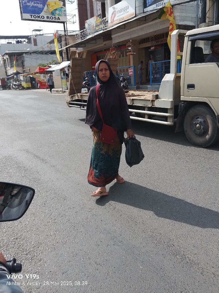 A woman wearing a black headscarf and a patterned skirt, and carrying a red handbag and a black plastic bag, is walking across a street in Jatibarang, Indonesia. She is walking in front of a white truck parked on the side of the road. The street is paved with asphalt and there are several shops on either side of the road, including one with the name "Toko Rosa" on a red sign. The woman's shadow is cast on the road in front of her. The image was taken on March 28, 2025, at 08:39.
