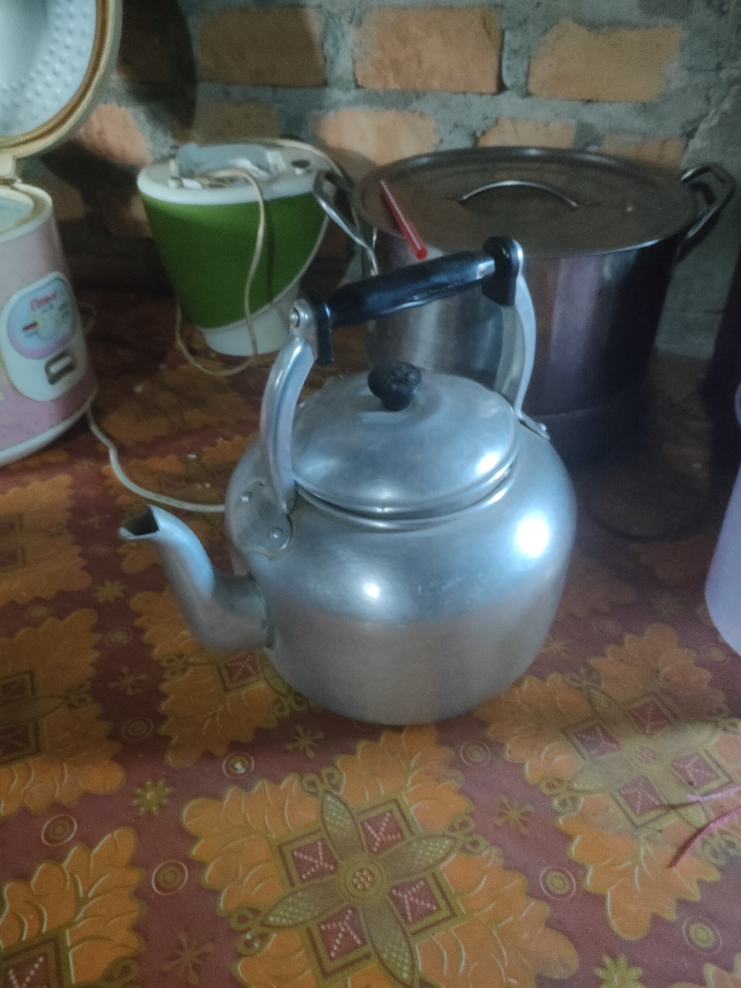 This is a close-up, eye-level, medium shot of a metal kettle in a kitchen setting. The kettle is made of brushed aluminum and has a black handle and a dark knob on the lid. It is placed on a patterned tablecloth with orange and brown floral designs. To the left of the kettle, a portion of a pink and white electric rice cooker is visible. Behind the kettle, to the right, is a large silver pot with a lid, and a red drinking straw is sticking out of it. Further to the right, a blurred purple object is visible. The background consists of a brick wall. The lighting appears to be artificial and somewhat dim, creating reflections on the metal surfaces. The overall impression is of a functional, albeit slightly cluttered, domestic kitchen. The image does not contain any information about the city of Padang Dalom, Indonesia.