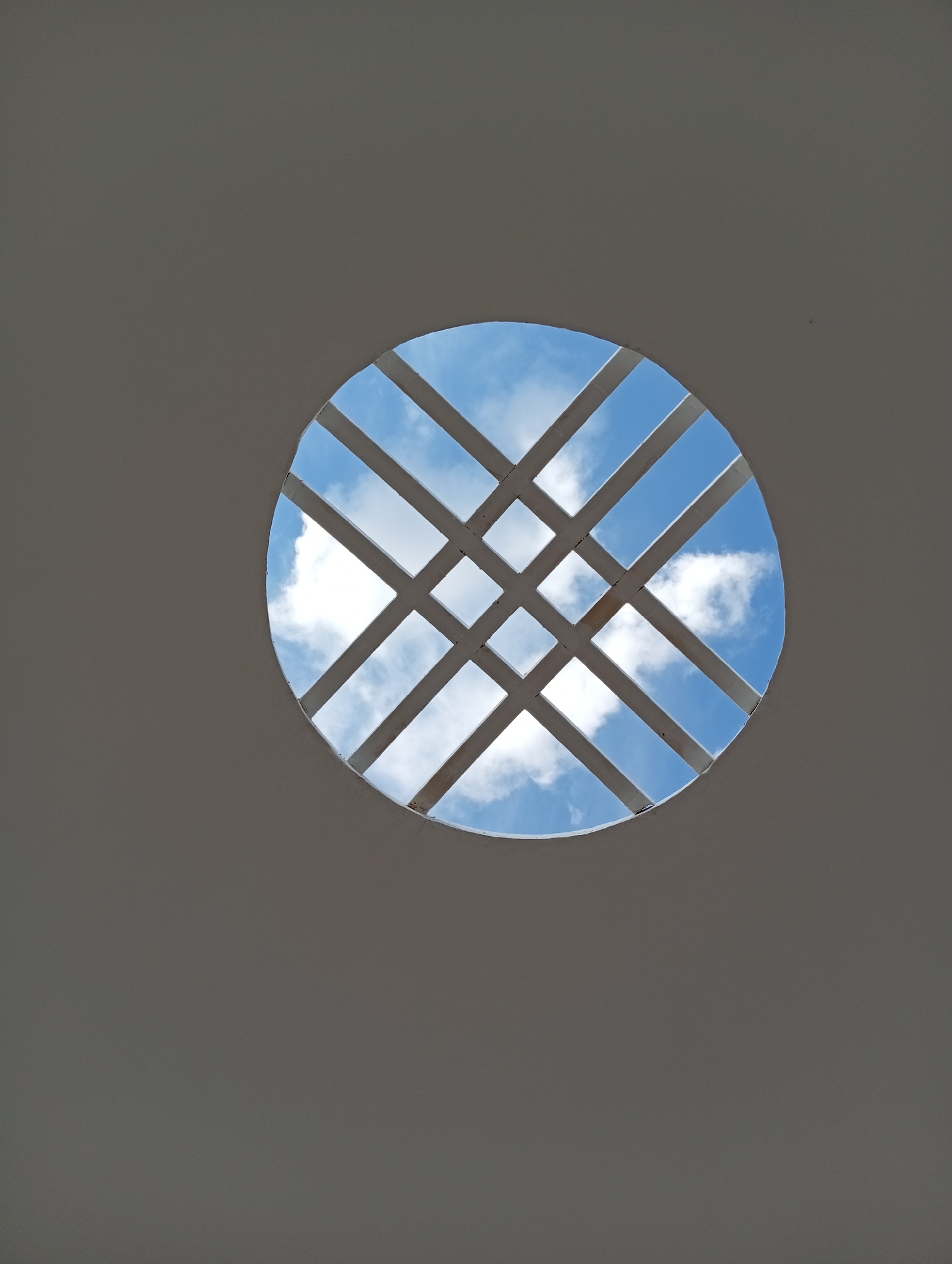 This image is a low-angle shot looking up at a circular skylight with a lattice of white beams. The beams form a diamond pattern, creating square and rectangular openings. Through these openings, a bright blue sky with scattered white clouds is visible. The surrounding structure appears to be a smooth, light gray surface, possibly a ceiling or dome. The image does not contain any discernible information about the city of Kostanay, Kazakhstan, or any people, actions, or interactions. It is a purely architectural and environmental observation.