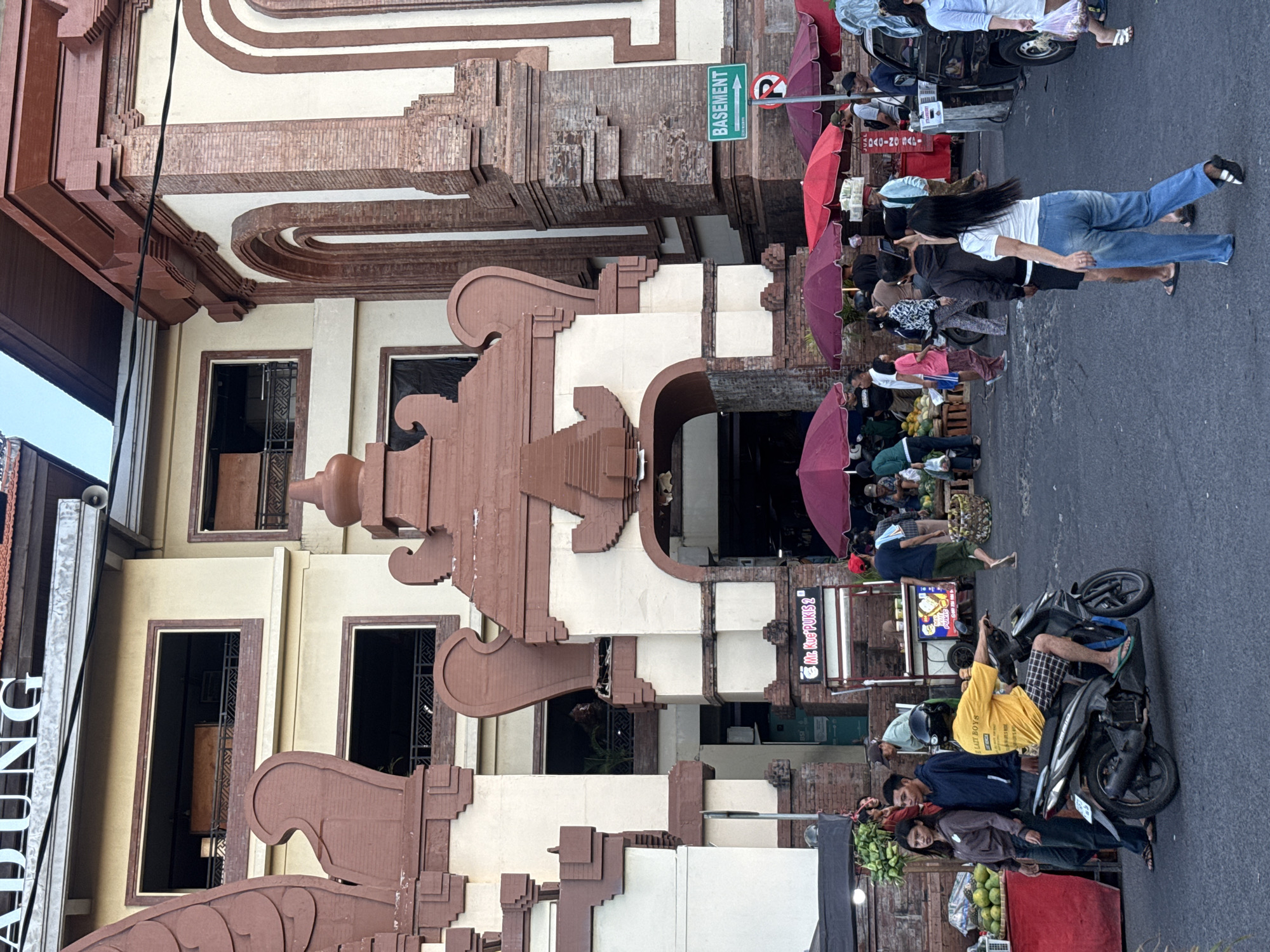 The image depicts a bustling street scene in front of a market in Denpasar, Indonesia. The architecture of the building features ornate, reddish-brown decorative elements and brickwork, with large arched entrances. A green sign with white lettering on the right side of the image reads "BASEMENT" with an arrow pointing to the right, and a circular red sign with a diagonal line indicates "no parking" or "no entry".
In the foreground, a man in a yellow shirt and plaid shorts is riding a dark-colored motorcycle. To his left, two young people, a woman and a man, are standing near a stall displaying green fruits, possibly coconuts or melons, on a red table. Further back, several food stalls are set up under red and purple umbrellas, with people browsing and interacting. Some stalls display signage, including one that says "Mr. KUE PUKIS 2". Various individuals are walking in different directions, some carrying bags. The ground is paved asphalt. In the background, the upper floors of the market building are visible, with open windows revealing balconies. The sky is clear and bright.