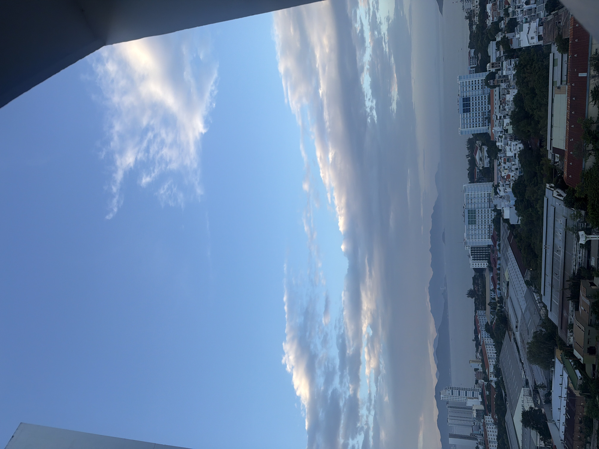 The image captures a panoramic view of Nha Trang, Vietnam, from a high vantage point. The sky is a vibrant blue, with scattered white and grey clouds. In the foreground, a white building structure partially frames the scene on the left and right. The middle ground reveals a dense urban landscape with a mix of modern high-rise buildings, residential structures, and industrial warehouses. Numerous rooftops, some with red or grey corrugated metal, are visible. The background showcases a serene body of water, likely the sea, with a distant mountain range silhouetted against the horizon. The overall atmosphere suggests a partly cloudy day with moderate visibility.