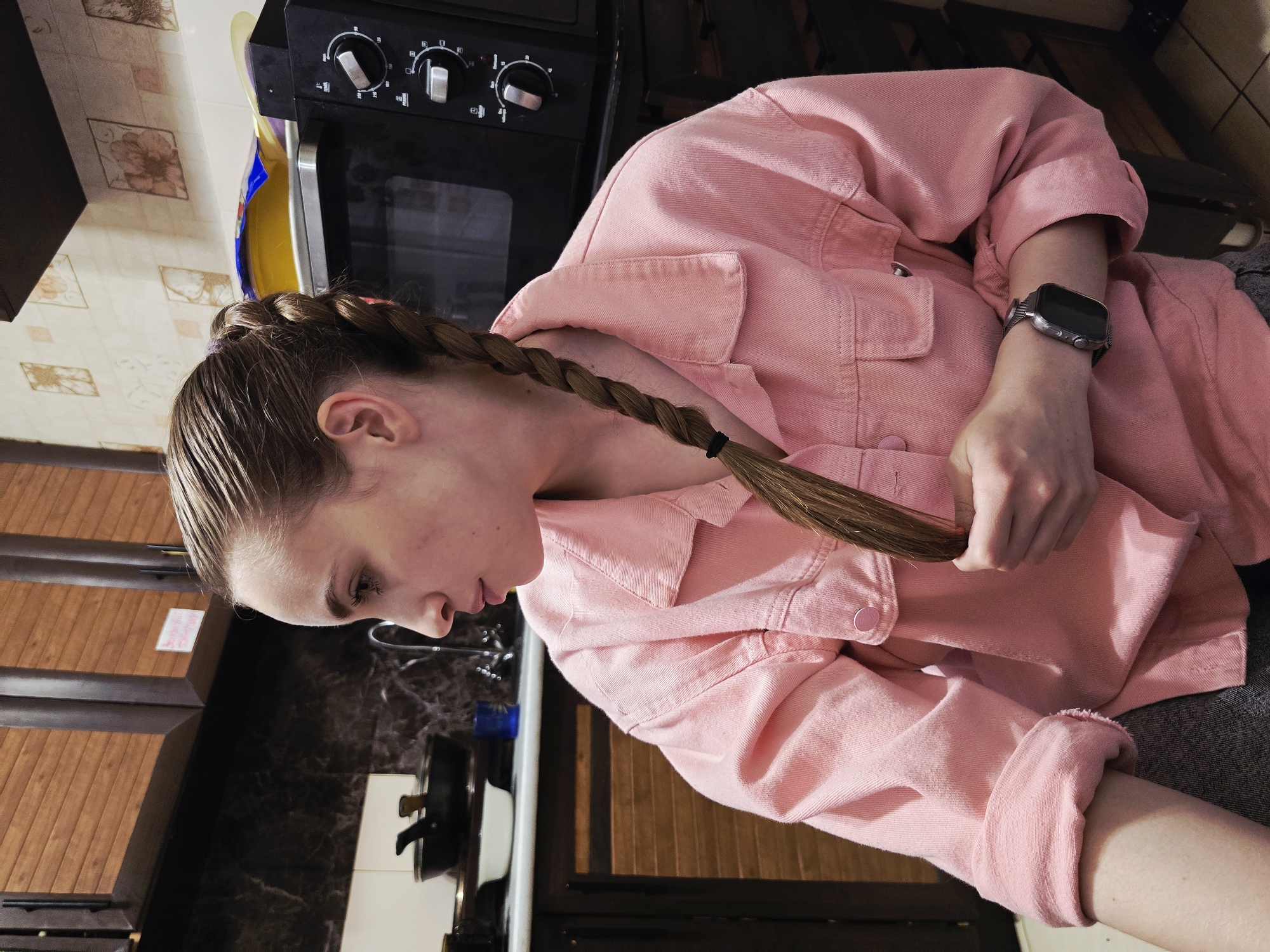 A young Caucasian woman with braided brown hair is seated in what appears to be a kitchen. She is wearing a pink denim jacket and a grey t-shirt. Her left arm is bent, and she is holding a strand of her braid with her fingers. A smartwatch is visible on her left wrist. In the background, kitchen cabinets, a microwave, and a countertop with various kitchen items are present. The lighting suggests it is indoors, possibly in the evening.