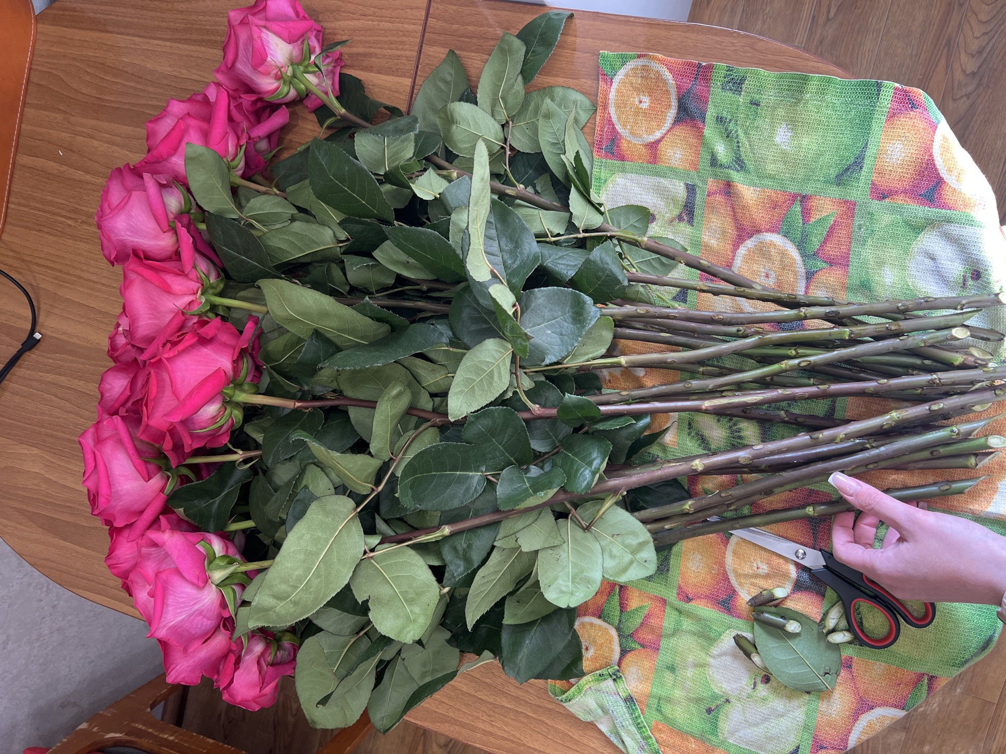 On a wooden table, a bouquet of pink roses is arranged with stems and leaves spread out across an orange-and-green patterned towel. A hand holds a pair of scissors, with snippets of stems and leaves lying nearby. The location is not discernible from the image.