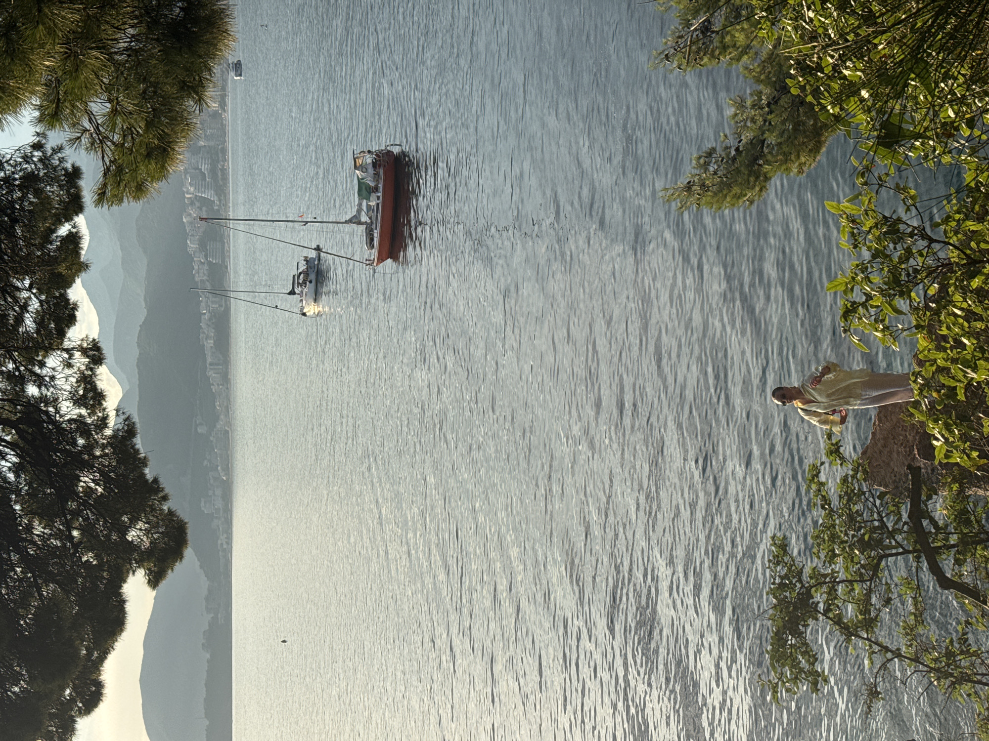 A vertical, eye-level, medium shot shows a woman in a light yellow outfit standing on a rocky outcrop covered in green foliage, looking out at the water. She is positioned in the lower right of the frame. The water is a deep blue with a rippled texture, reflecting the soft light of what appears to be a late afternoon or early morning sun. In the middle ground, two sailboats are anchored on the calm sea. The larger sailboat, in an orange hue, is closer to the foreground, and a white sailboat is further to the left. In the background, a coastal town with buildings is visible along a mountainous shoreline. The sky is a muted blue with a hint of haze. The scene is framed by the dark green needles of pine trees in the foreground, creating a natural border for the composition. The overall impression is one of serenity and natural beauty, with a human element present in the scene.