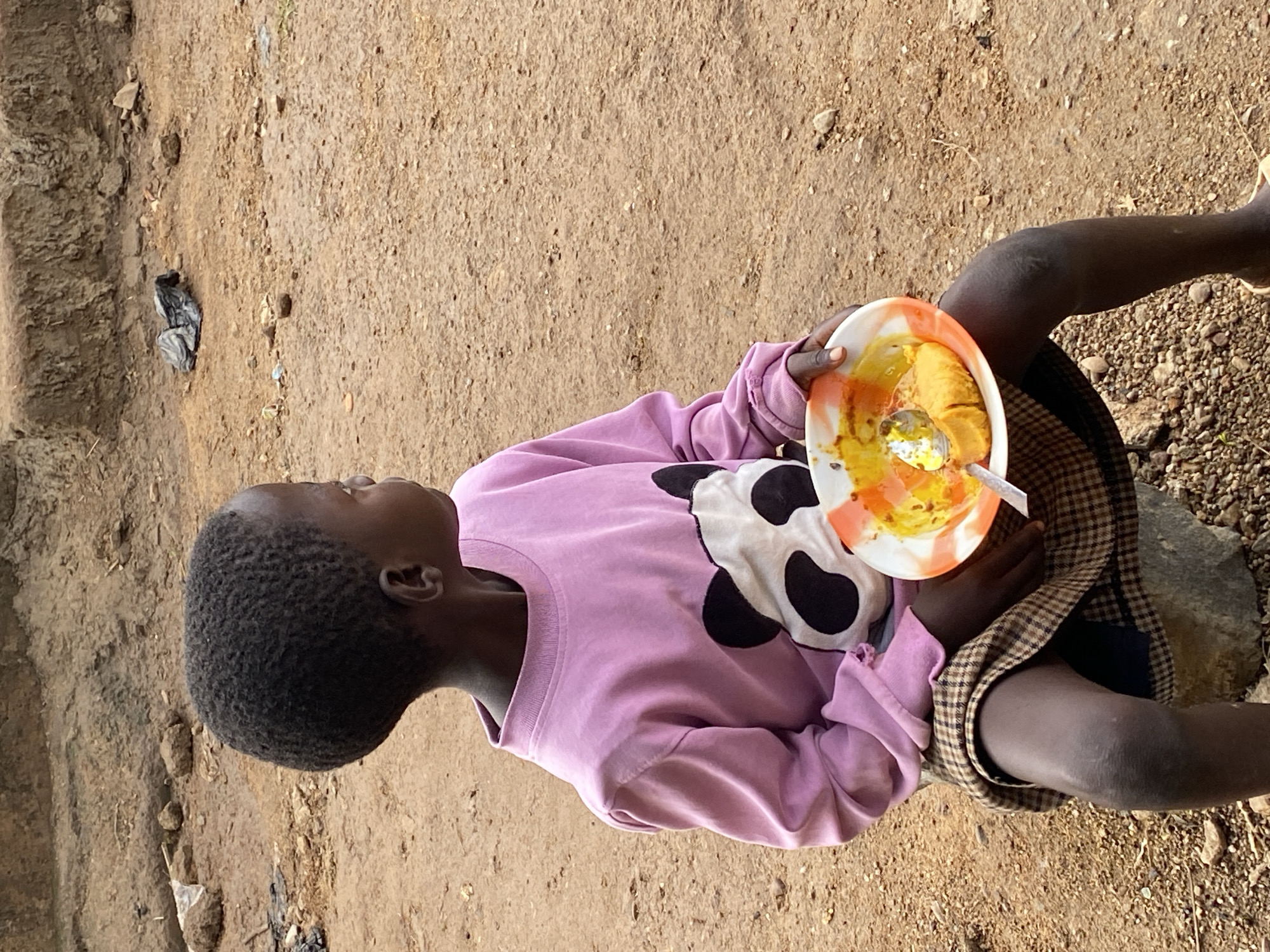 An overhead shot captures a young child holding a plate of food outdoors in Riyom, Nigeria. The child, with dark skin and short, kinky black hair, is seen in profile, looking upwards or straight ahead. They are wearing a long-sleeved, light pink sweatshirt featuring a black and white cow-print design on the front, and a plaid skirt in shades of brown and black.
The child is seated on an unseen surface, possibly a low stone, with their legs bent and visible. In their hands, they hold a round plastic plate with an orange and white swirl pattern, containing a yellowish-orange meal, likely a thick porridge or stew. A silver spoon is visible in the food.
The setting is an unpaved, outdoor area characterized by dry, light brown soil or sand mixed with numerous small rocks and pebbles. To the left, a slightly darker, textured earthen bank or wall is visible. The natural lighting suggests it is daytime, with soft shadows indicating either early morning or late afternoon. The environment appears arid and rural or semi-urban. No discernible text is visible in the image, and the child's expression is neutral, suggesting focus on the meal.
