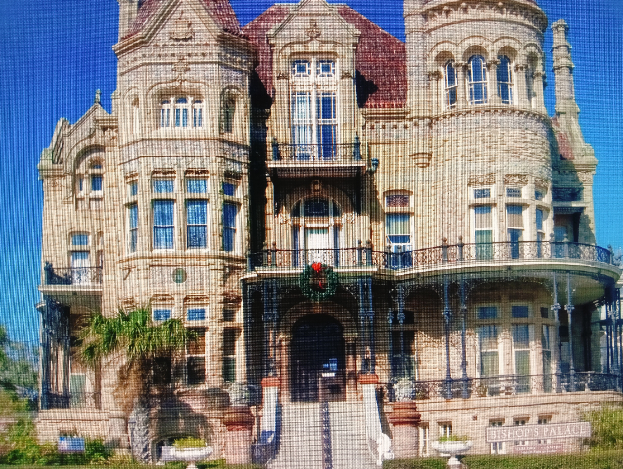 The image shows a large, ornate, stone mansion under a clear blue sky. The architecture is elaborate, with multiple towers, arched windows, balconies with decorative ironwork, and a red tiled roof. A wreath with a red bow is hanging above the main entrance, suggesting a holiday season. A palm tree is visible on the left side of the building, and manicured bushes and urns are present in the foreground. A sign in front of the building reads "BISHOP'S PALACE." This building is Bishop's Palace in Galveston, Texas, USA, not in Sluck, Belarus. There are no people visible in the image.