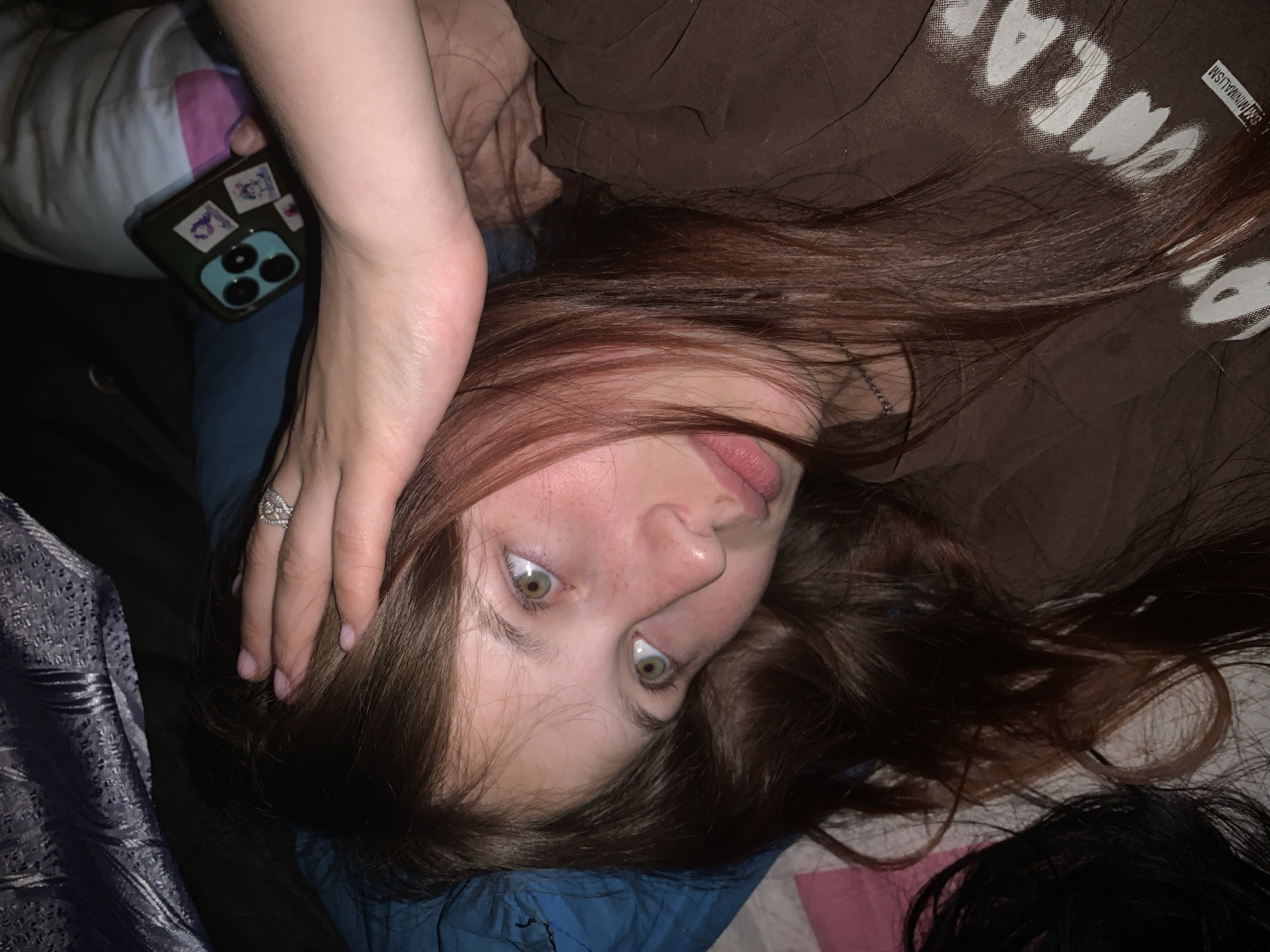 A young woman with long auburn hair is lying down, looking up at the camera. Her left hand rests on her forehead, and she is wearing a ring. She has fair skin, freckles, and green eyes. She is wearing a brown shirt with white text. To her left, a phone with stickers is on a pillow with a pink and white pattern. The background includes a blue blanket and a gray patterned fabric. The location, Фоки, Russia is not apparent from the image.