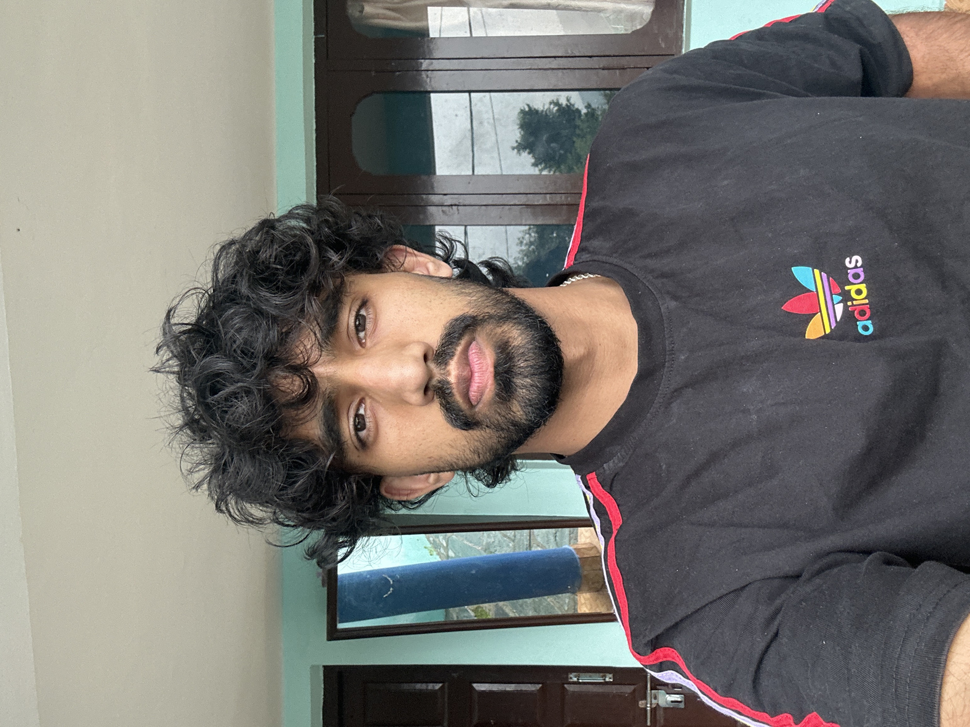 A young man with curly dark hair, a beard, and a light complexion is pictured from the chest up, facing the camera. He is wearing a black t-shirt with a colorful Adidas logo on the left side of his chest and red and white stripes on the sleeves. The background appears to be an outdoor setting, possibly a porch or balcony, with a light blue wall and a brown door visible on the left. A window with dark brown frames and curtains is visible on the right. A mirror is also present on the left, reflecting a blue cylindrical object and a sliver of the man's face.