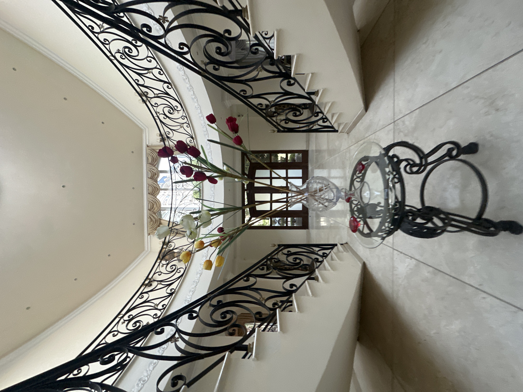 The image depicts the interior of a building, potentially a residence, located in Jesús del Monte, Mexico. The prominent features are a curved staircase with elaborate black wrought-iron railings and a glass table adorned with artificial flower arrangements. The staircase, with white risers and treads, ascends towards a window or glazed door, partially visible in the background. The flooring appears to be light-colored tile or stone. The color palette is dominated by neutral tones, contrasted by the black ironwork. No individuals are visible in the scene.