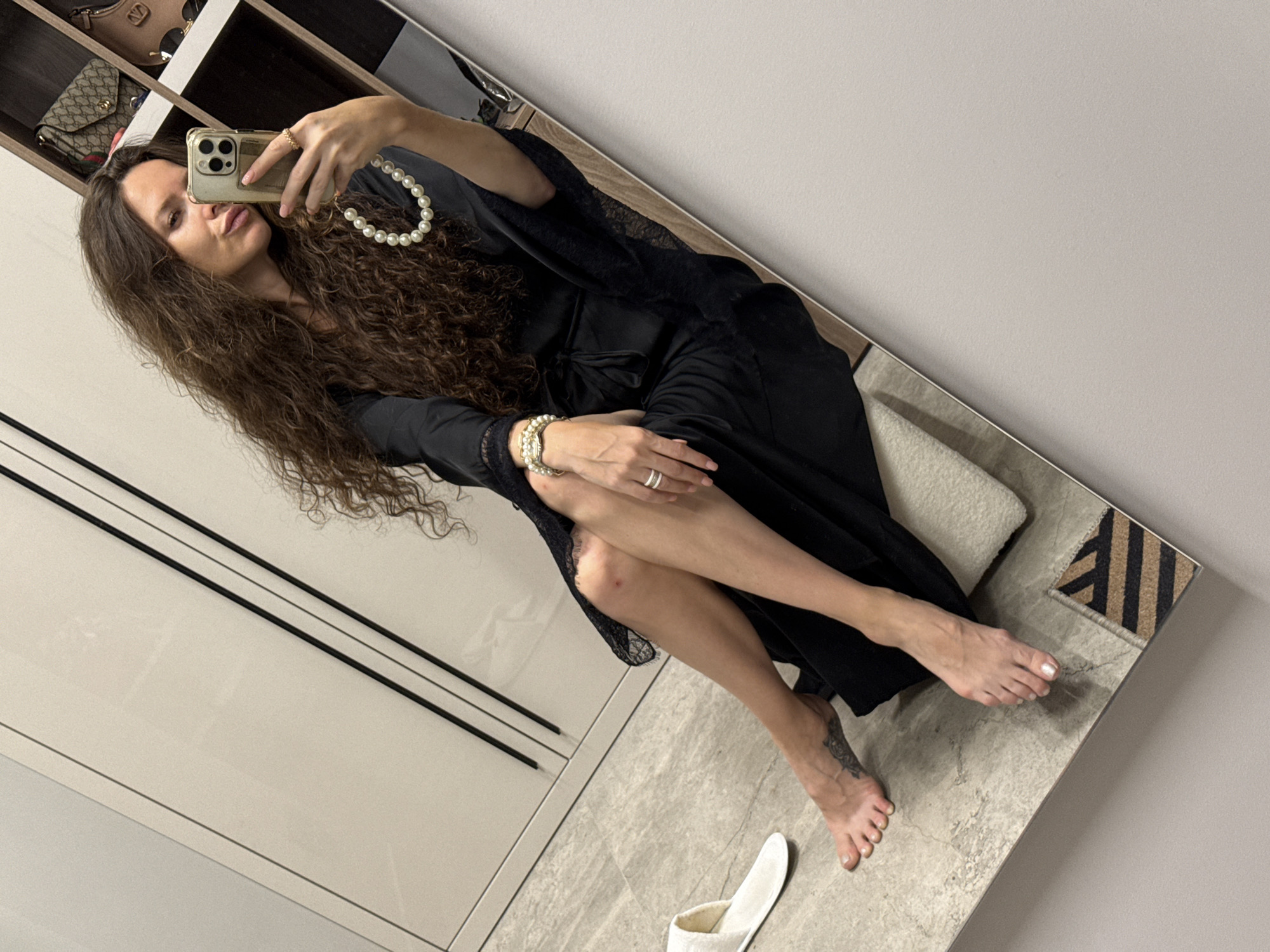 In this media file, a woman with long, curly brown hair is captured in a mirror selfie. She is wearing a black negligee with lace trim and a pearl necklace. She is also wearing bracelets and rings on her hands. Her left leg is crossed over her right, and her feet are bare. A tattoo is visible on her left ankle. In the background, a closet with shelves containing handbags and a mirror are visible. A white slipper is on the floor in front of the mirror. The lighting suggests it is daytime. The woman's facial expression is playful, with her lips pursed.