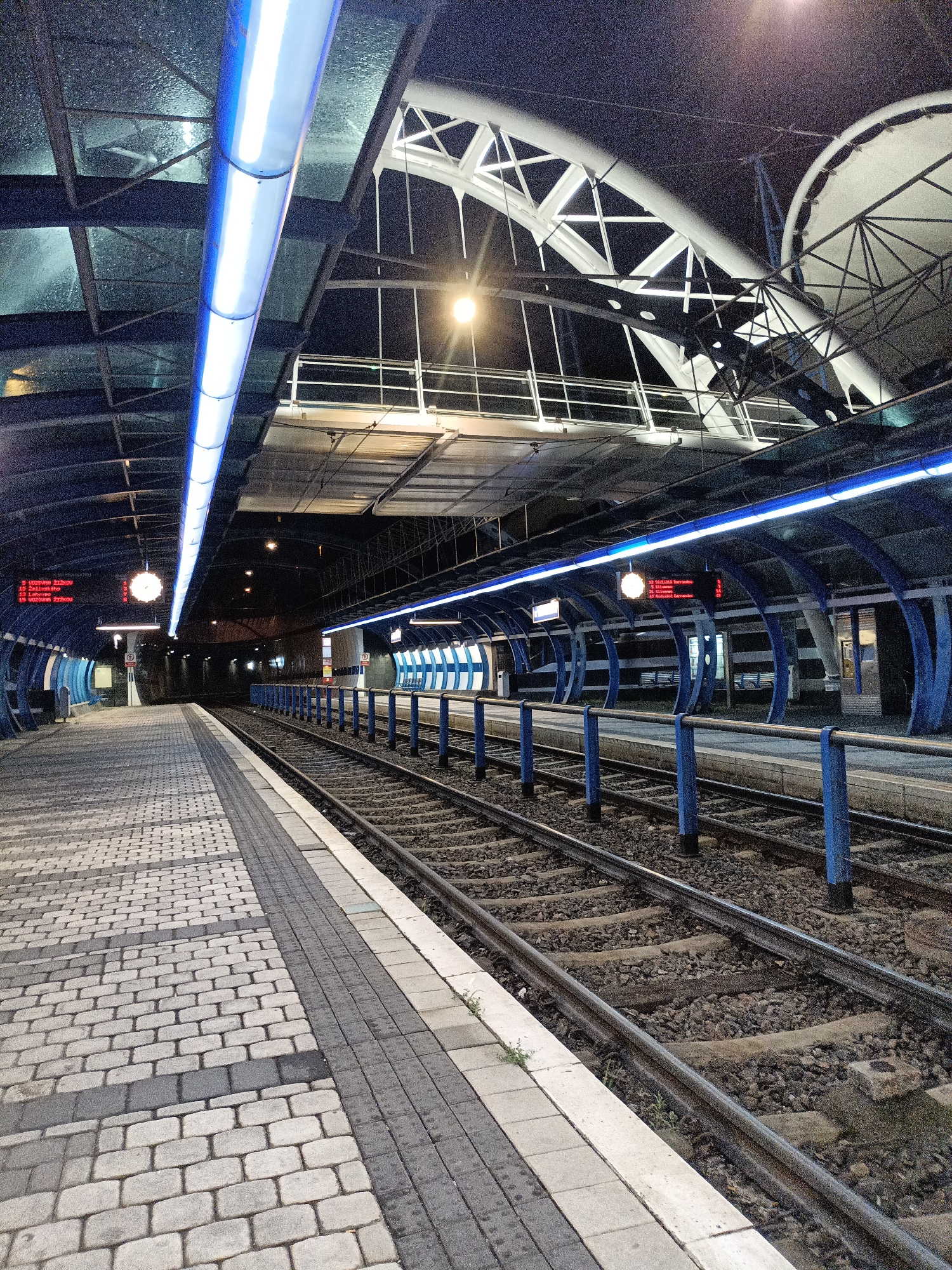 The image depicts a modern, empty train station platform at night. The platform is paved with cobblestones and bordered by train tracks. A series of blue pillars supports a curved roof structure, illuminated by long, bright blue fluorescent lights. Above, a white, arched canopy with structural elements is visible against the dark night sky. Digital displays show information, and a clock hangs from the ceiling. The station appears to be part of the Prague Metro system, specifically possibly a station like Náměstí Míru or Florenc, which feature contemporary architectural designs with extensive use of glass, metal, and modern lighting. There are no people or vehicles visible in the scene.