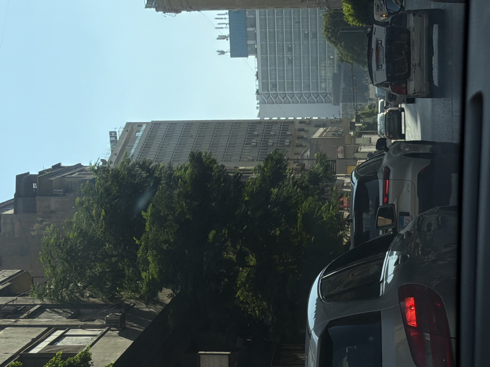 The image depicts a traffic scene in Cairo, Egypt, likely on a street lined with multi-story buildings. In the foreground, the rear of a silver car is visible, its brake lights illuminated. Several other cars are in traffic, including a white SUV and a silver sedan further down the road. Some cars appear to have their trunks open, with items visible inside. Tall, weathered buildings with visible windows and air conditioning units line the left side of the street, partially obscuring a dense cluster of green trees. In the background, larger, modern buildings with glass facades and what appear to be communication antennas on their roofs can be seen under a clear blue sky. The overall impression is a busy urban environment.