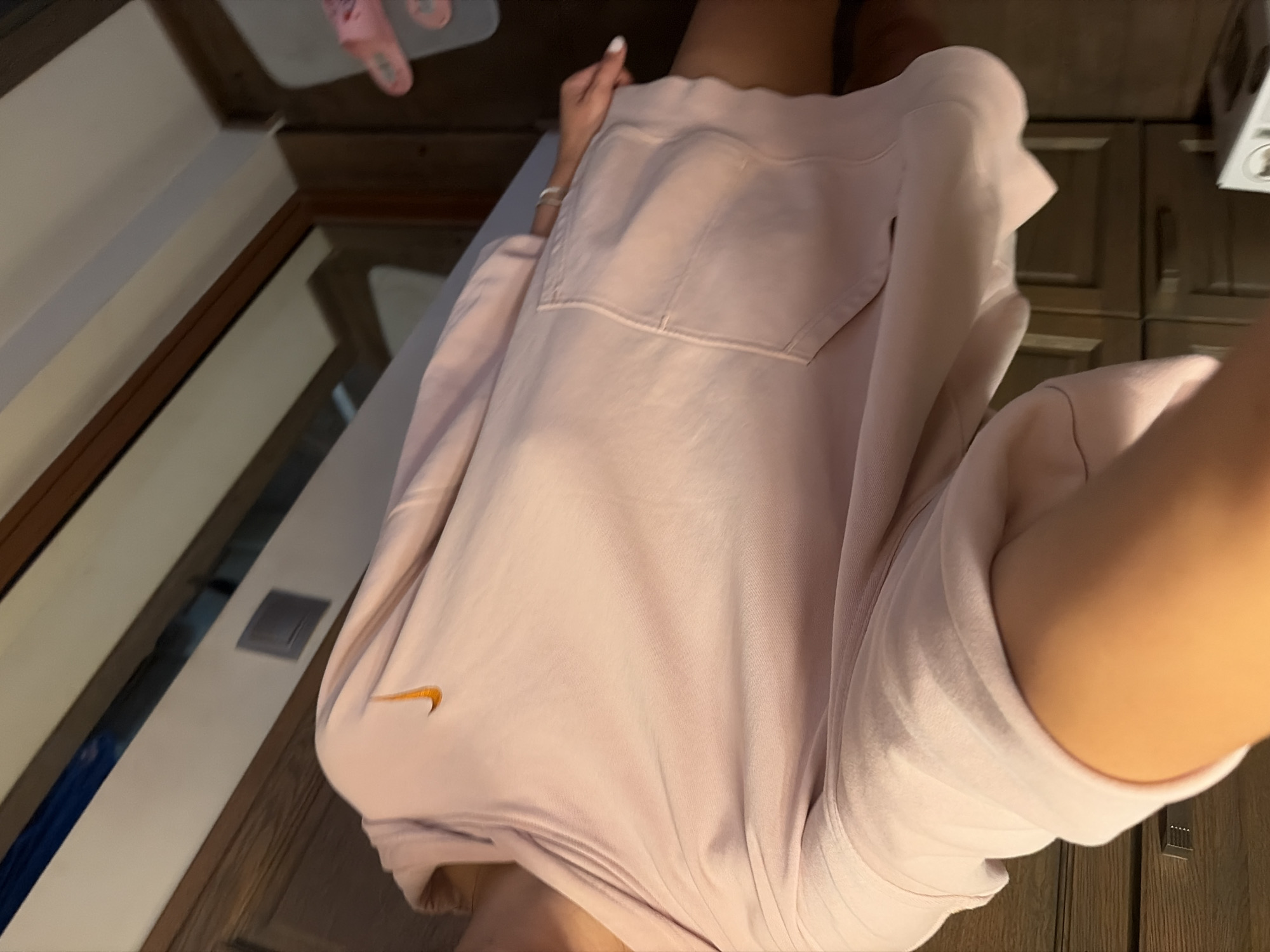 A person is shown from the chest down, wearing a light pink Nike hoodie. The hoodie has a pocket on the right side and the Nike logo is visible on the left breast. The person is standing in a room with wooden furniture, including cabinets and drawers. There are pink slippers visible in the background. The room appears to be a bedroom or dressing area. The image is taken from a low angle, looking upwards. The lighting is dim, suggesting it is nighttime or the room is not well-lit. The specific location in Tsarevo, Bulgaria cannot be determined from the image alone.