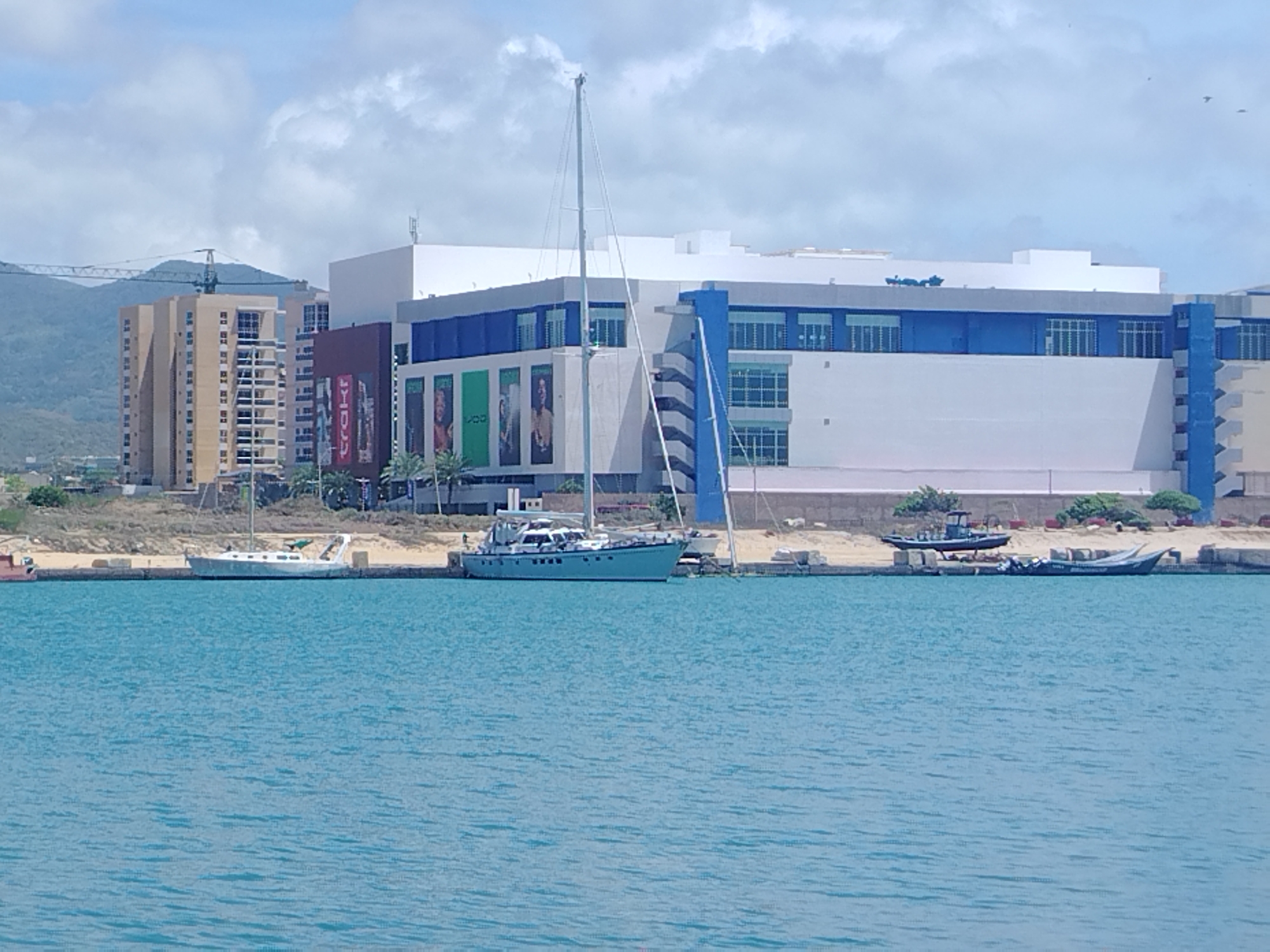 The image depicts a waterfront scene in Porlamar, Venezuela, under a bright, partly cloudy sky, suggesting daytime. In the foreground, a calm, turquoise body of water dominates the lower half of the frame. Moored along a concrete barrier in the mid-ground are several boats.
A large white sailboat with a tall mast is prominently positioned, its hull resting on the shore. To its left, a lighter-colored motorboat is also beached. Further to the right, a dark-colored boat with a small cabin and a few outboard motors is visible, as well as another lighter-colored boat partially obscured by it.
In the background, a modern complex of buildings stands. The most striking is a large, multi-story structure with sections of white and bright blue facades. Large windows and balconies are visible on some parts of the building. To the left of this main structure, a taller, tan-colored residential building with numerous balconies is situated, with a construction crane visible behind it. The landscape behind the buildings appears to be hilly and somewhat arid, with some vegetation.
The scene conveys a sense of a coastal town or city, possibly a harbor or marina area, with ongoing construction or development evident. The weather appears pleasant, conducive to outdoor activities on the water. No people are clearly discernible in the image, but the presence of boats suggests maritime activity. Some faint text is visible on the posters on the side of the building, but it is not legible.