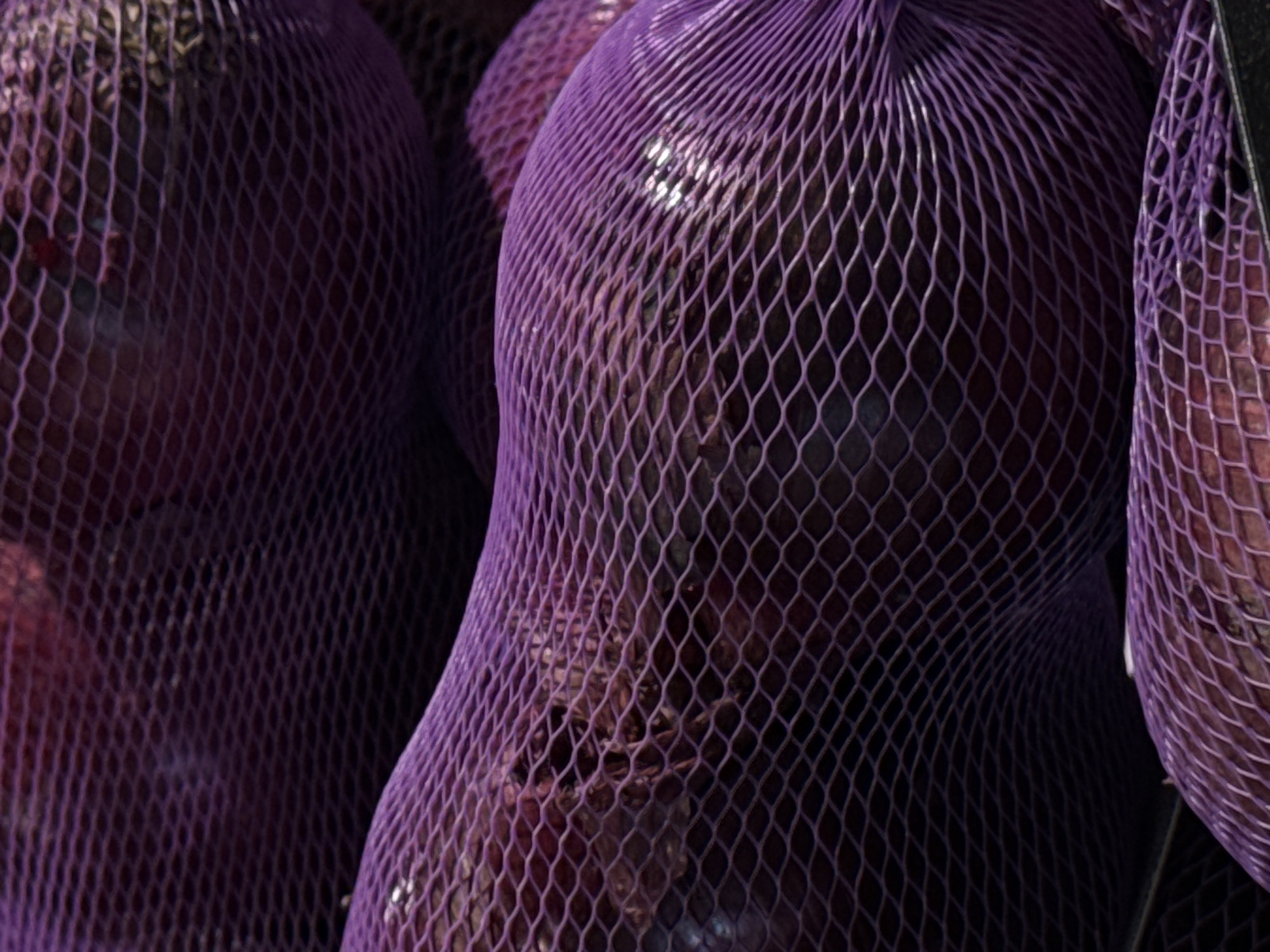 The image is a close-up, high-angle shot of several purple net bags filled with what appear to be onions or similar produce. The bags are tightly packed and create a textured surface with a diamond-shaped mesh pattern. The lighting is dim and casts subtle shadows, highlighting the rounded shapes of the produce within the bags. The overall color palette is dominated by deep purples and dark tones, with some brighter highlights reflecting off the surface of the produce. The scene appears to be indoors, likely in a market or storage area. There are no people visible in the image. The image is purely descriptive and does not contain any information about its location in Moscow, Russia, or any other specific geographical details.