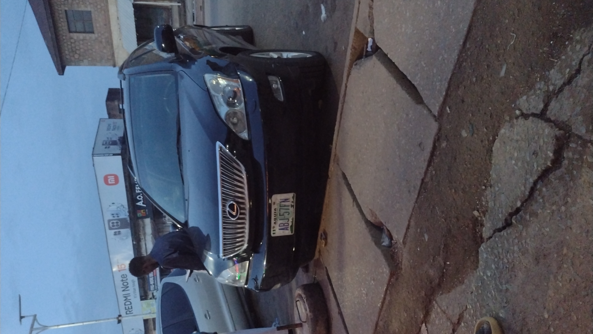The image captures a street scene, likely at dusk or early morning given the soft, ambient light and pale blue sky. The primary subject is a dark-colored Lexus SUV, positioned on what appears to be a cracked and uneven paved surface next to a darker, possibly dirt or gravel, shoulder. A person, identified as a dark-skinned male wearing a blue top, is partially visible leaning into the driver's side of the Lexus, suggesting interaction with the vehicle such as entering, exiting, or adjusting something inside. Another white car is partially visible to the left of the Lexus.
The Lexus bears a Nigerian license plate, clearly displaying "ABUJA" and the alphanumeric sequence "ABJ-57FN". In the background, a large billboard is visible with the text "REDMI Note 15" and the "mi" (Xiaomi) logo, along with partially visible text "A.O. FF...". Behind the billboard, a building with stone-faced walls and a single window can be seen. The ground surface features significant cracks and varying textures of concrete and dirt. A street light pole is also visible on the far left. The overall setting points to an urban or semi-urban area in Nigeria, consistent with the provided context of Jos. No specific weather conditions are evident.