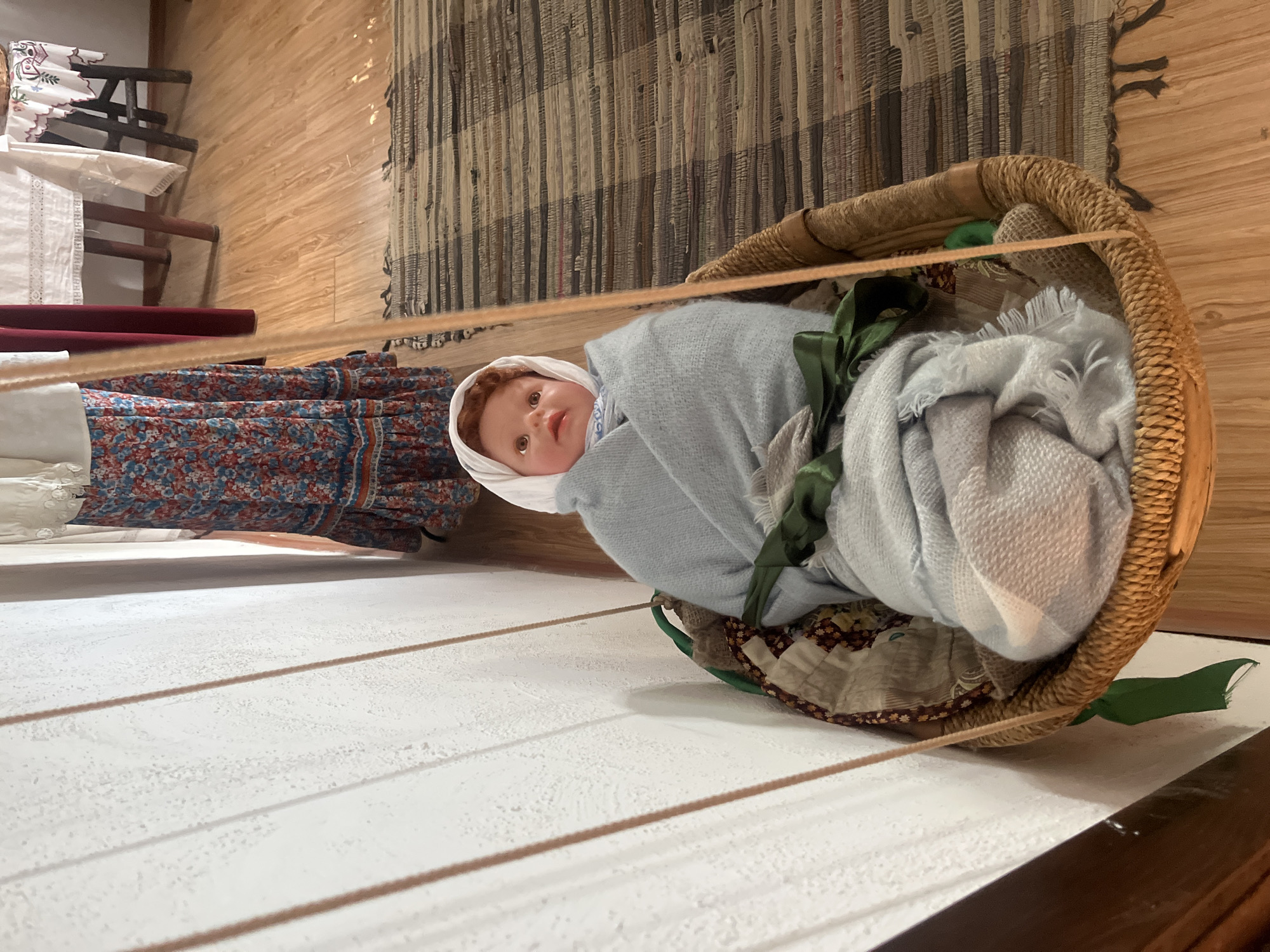A doll representing an infant is cradled in a woven basket swing, suspended by ropes attached to a white wall. The doll is wrapped in a pale blue blanket, secured with a green ribbon. Beneath the blanket, a patterned quilted fabric is visible. The swing rests on a striped rug with fringe, laid on a wooden floor. In the background, a table draped with a white tablecloth and a partially visible dress with a floral pattern suggest an interior setting. The overall scene evokes a domestic or historical exhibit, possibly within the Taman Local History Museum, given the context of the city.