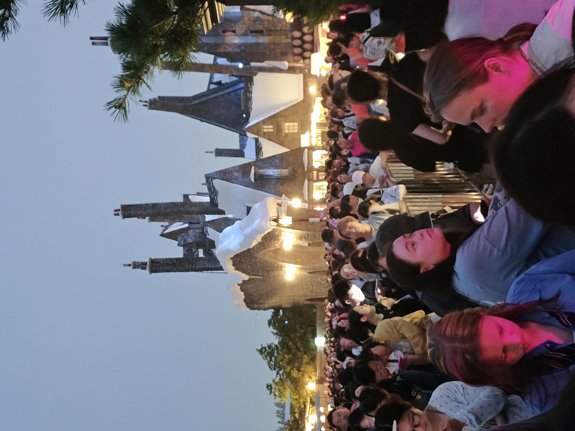 The image depicts a crowd of people gathered outdoors, seemingly at a nighttime event or attraction. The setting appears to be a themed area with buildings that resemble old-fashioned structures with stone chimneys and snow-dusted roofs, reminiscent of a fantasy or historical village. String lights are visible, casting a warm glow on the scene and illuminating the faces of the people.
The people are a diverse group, with many of them looking upwards, suggesting they are watching a performance, a light show, or some form of entertainment. The mood seems to be one of anticipation and engagement. Some individuals are illuminated by pink light, adding a dramatic effect to the scene. The sky is dark, indicating that it is likely evening or night.
Notable details include the architectural style of the buildings, the presence of greenery suggesting trees, and the way the crowd is densely packed. The pink lighting is a prominent visual element, highlighting some of the faces and creating a vibrant atmosphere. There is no visible text in the image. The location context provided is Tongzhou District, China, which might imply this is an attraction within that district.