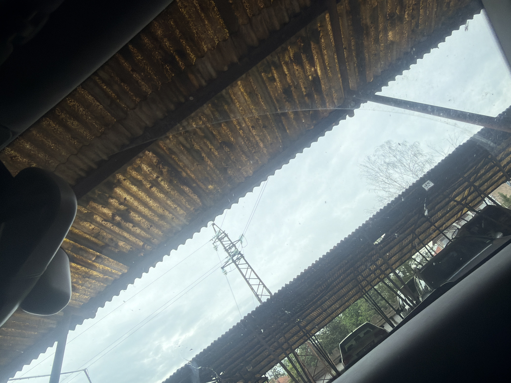 The image is captured from inside a vehicle, looking upwards and slightly forward through its windshield or sunroof. Parts of the vehicle's dark interior, possibly the headliner or sun visors, frame the top and bottom right edges of the view.
The central and most prominent feature is a vast, weathered corrugated canopy or roof structure, seen from underneath. It consists of translucent, yellowish-brown corrugated sheets, likely fiberglass or plastic, supported by dark metal framing. This structure appears old and dirty, suggesting an aged carport, a covered parking area, or an open-air workshop.
Through the large openings in the corrugated roof and the vehicle's glass, an overcast sky is visible, indicating a cloudy daytime. Against this backdrop, a tall, metal lattice electricity pylon with power lines is clearly visible on the left side of the frame. Sparse, bare tree branches are also silhouetted against the sky, suggesting a cold season like late autumn or winter.
In the lower right background, under the corrugated canopy, several other vehicles (cars) are partially visible, suggesting a parking or storage area. Some green foliage is also present further in the background. A small, unreadable white rectangular sticker is affixed to the vehicle's glass near the top right, and another small, unreadable sign is visible on a structure in the distant background on the right.
The scene depicts a static, daytime view of an outdoor, utilitarian area, consistent with a setting in Dolna Mitropolia, Bulgaria. There are no people visible, and no specific activity or event is taking place. The weather is cloudy.