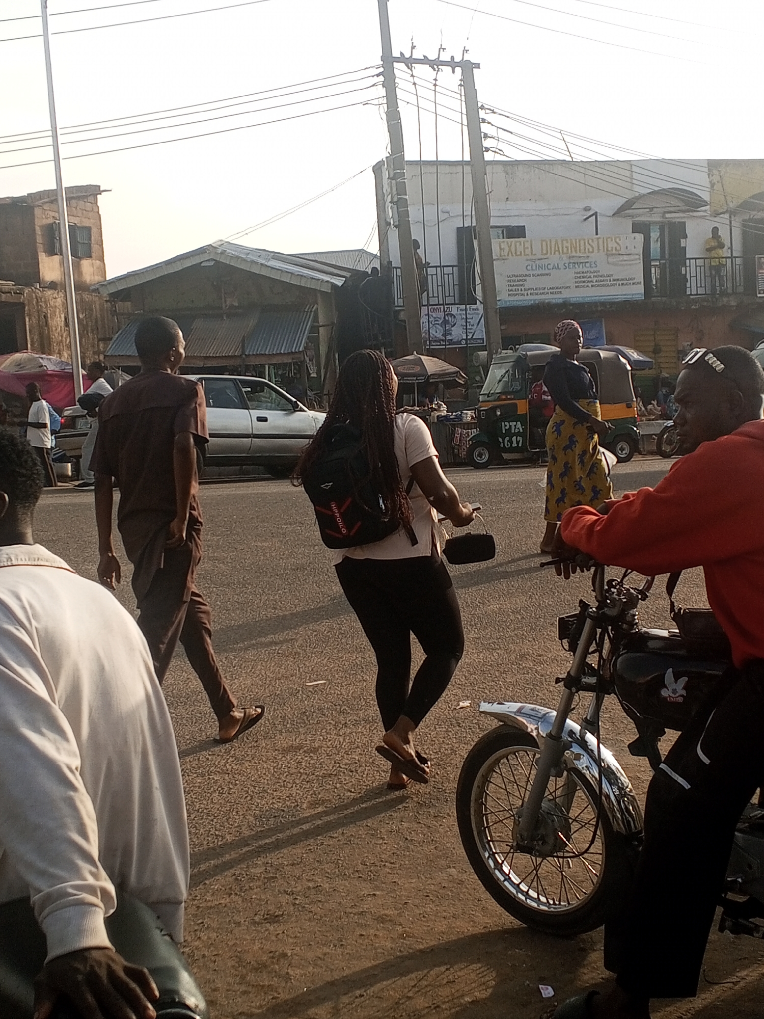 The image depicts a street scene in Gwong, Nigeria. A man in a brown suit walks left to right across the frame, and a woman with long braids, wearing a white shirt and black leggings, walks perpendicular to him. Both are pedestrians. A man in a red shirt sits on a parked motorcycle to the right. A woman in a patterned dress stands near a three-wheeled vehicle ("Keke") marked "PTA 1617". In the background, there is a building with "EXCEL DIAGNOSTICS" sign visible. Other people, vehicles, and roadside stalls are visible in the periphery. Electrical poles and wires are above the scene.