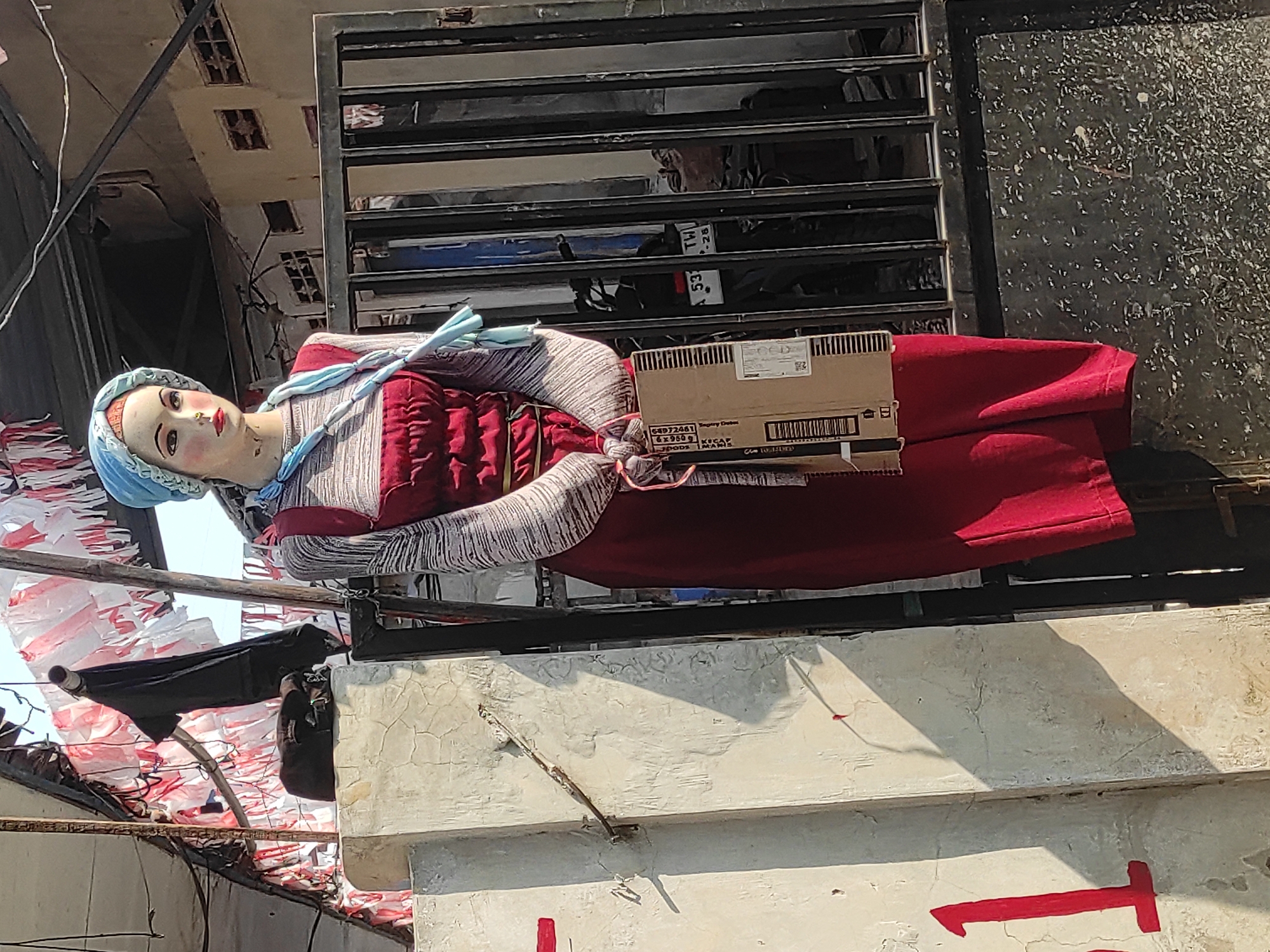 A full-body mannequin is displayed outdoors, positioned in front of a black metal gate and a rough, light-colored concrete wall. The mannequin wears a blue headscarf, a striped long-sleeved shirt, and a red dress with suspenders. It is holding a cardboard box. The background includes a motorcycle parked behind the gate and a building with windows. Red and white streamers are strung across an overhead structure. A red numeral "1" is painted on the concrete wall. The scene is illuminated by sunlight, casting shadows. The location is identified as Cankuang, Indonesia.