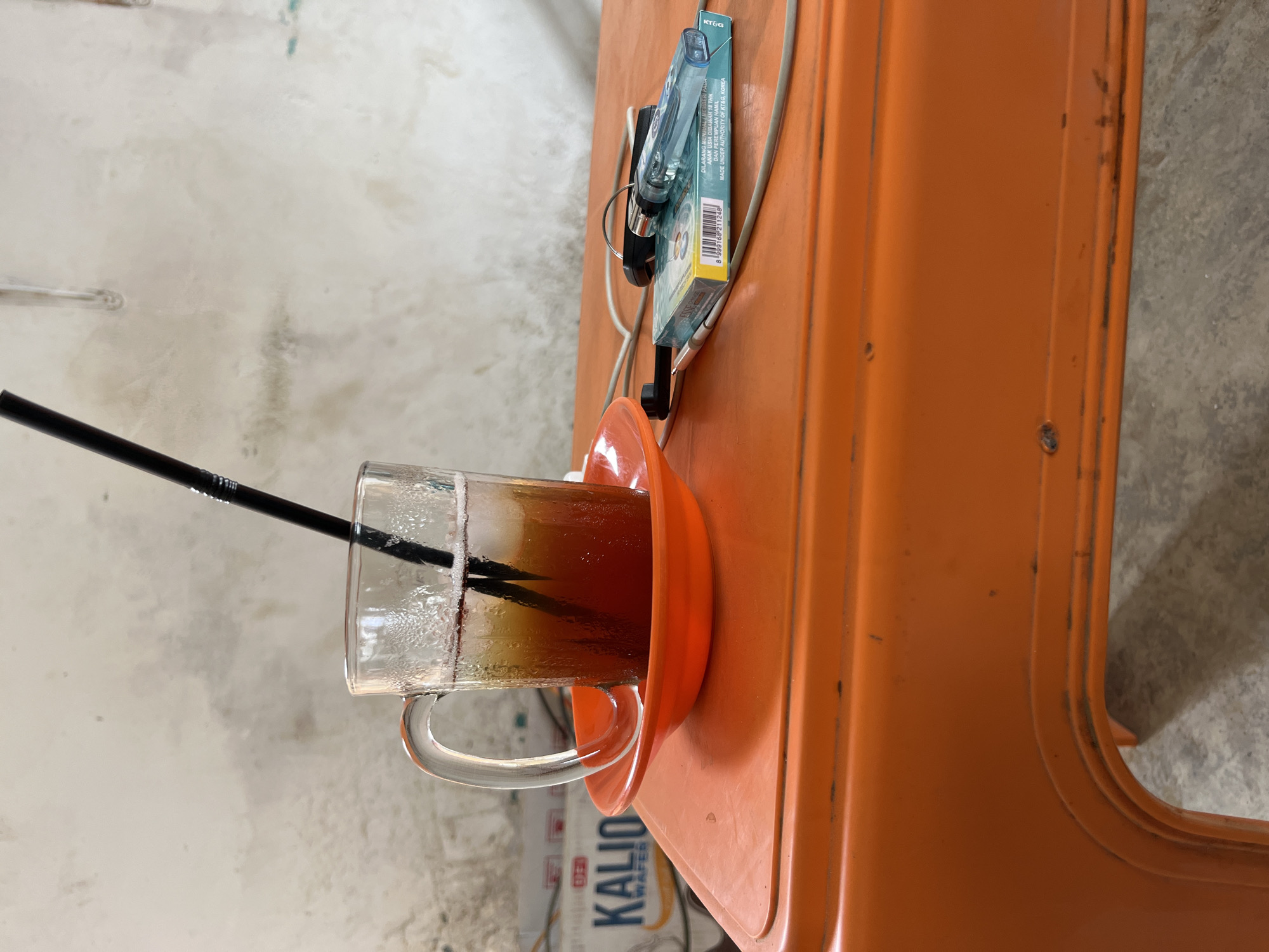 A glass of iced tea with a black straw sits on an orange saucer, which is on an orange table. Next to the glass are a pack of cigarettes, a lighter, and keys. In the background, a white wall is visible. The scene is illuminated by natural light.