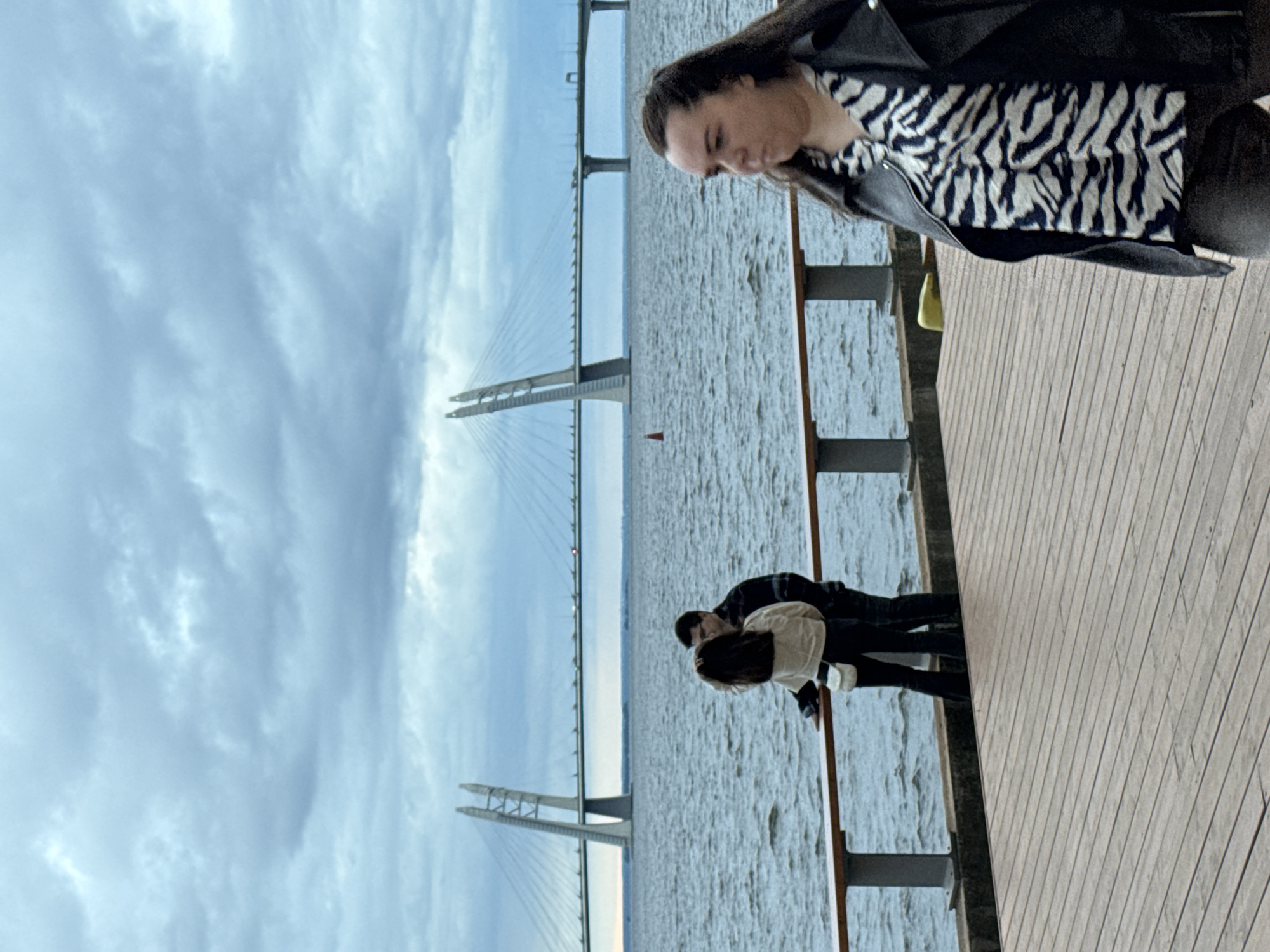 This image captures a cloudy day scene on a wooden boardwalk beside a body of water in Saint Petersburg, Russia. In the foreground, a woman with dark hair is walking away from the camera, wearing a black leather jacket over a black and white zebra-print shirt and dark pants. In the middle ground, a couple stands by a railing, facing away from the camera and towards the water. The woman has dark hair and is wearing a light-colored top and dark pants, with a small white bag slung across her body. The man beside her has dark hair and is wearing a dark plaid shirt. In the background, a wide cable-stayed bridge spans across the water under a heavy, overcast sky. The bridge's distinctive A-shaped pylons are visible. A small red buoy floats in the water to the left of the bridge. The boardwalk is constructed of parallel wooden planks, and a metal railing with vertical supports lines the edge of the water.