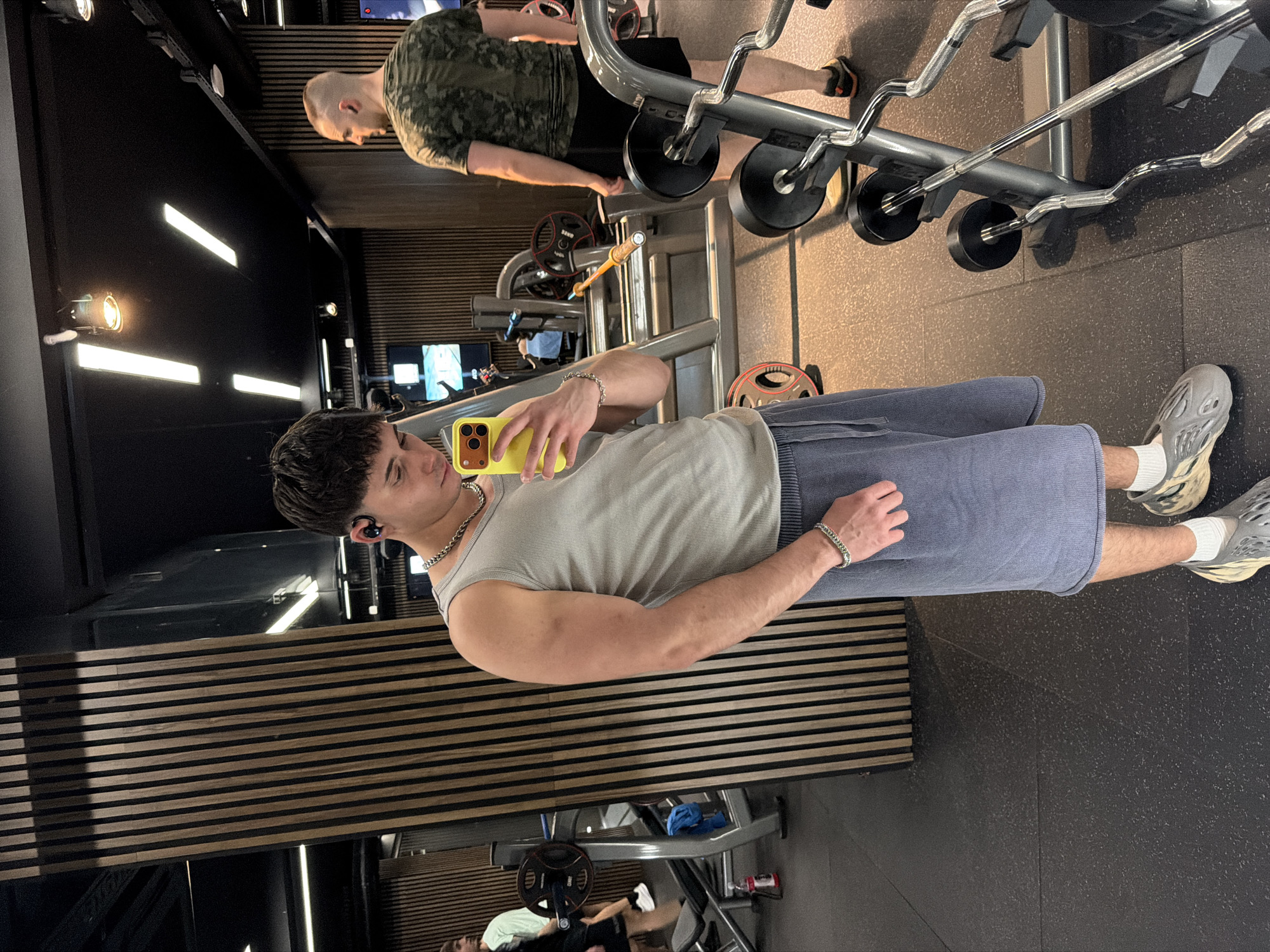 The image depicts an indoor gym located in Sofia, Bulgaria. The primary subject, a male in his late teens or early twenties with dark, short hair, is positioned slightly left of center, looking down at a yellow-cased smartphone he holds in his right hand, seemingly capturing a self-portrait. He wears a light grey ribbed tank top, blueish-grey sweat shorts, white socks, and grey textured slip-on athletic shoes. He is accessorized with black earbuds, a silver chain necklace, and a silver chain bracelet on his left wrist. His physique indicates muscular development, particularly in his arms.
In the mid-ground to the right, another male with a closely shaved head is visible from the back, wearing a camouflage-patterned t-shirt, black shorts, and dark athletic shoes with orange accents. He stands facing right. A partial reflection of a third male is visible on the far left, wearing a light-colored t-shirt and dark shorts.
The gym environment features a dark, speckled rubberized floor. Wall elements include prominent vertical wooden slats set against dark panels. The ceiling is dark, equipped with modern lighting fixtures comprising linear white LED strips and warm-toned circular spotlights. Various gym equipment is present, including barbells, weight plates, and a barbell storage rack on the right. A red water bottle is on the floor to the left.