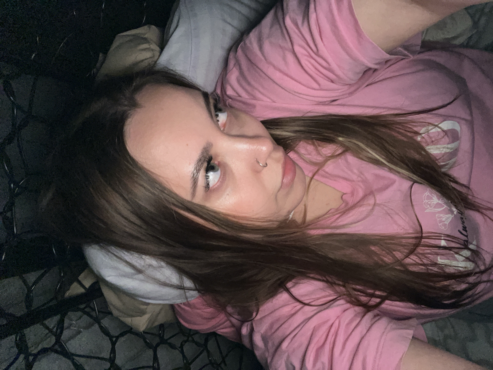 A young woman with long brown hair is lying down, looking upwards towards the camera. She has fair skin, light eyes, and a nose piercing. She is wearing a pink t-shirt and appears to be resting on a pillow. The background is dark and features what looks like a metal headboard or frame. The specific location in Novopokrovskaya, Russia is indeterminable from the image.
