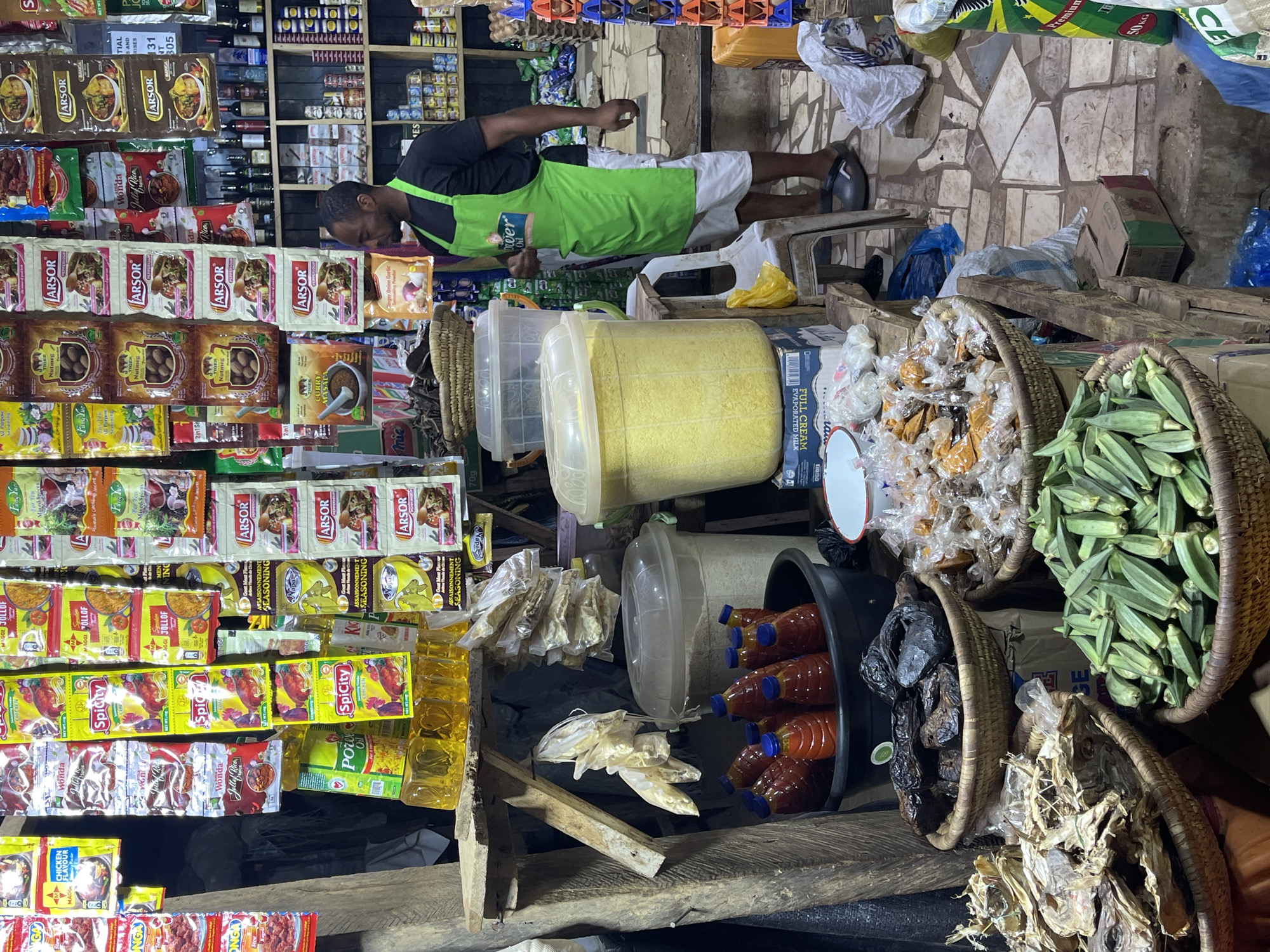 This image captures a bustling indoor market stall in Abuja, Nigeria, filled with a diverse array of food products and ingredients. A man, likely a vendor, is depicted in the center, wearing a black t-shirt, white shorts, and a bright green apron. He is leaning forward, seemingly attending to or arranging the merchandise.
The stall is densely packed with goods. Shelves are lined with numerous small sachets and packets of spices and seasonings, including prominent labels such as "ARSOR," "MAGGI JOLLOF," "Spicity," "Wonda Jollof Rice," "TIGER Nutmeg," "TIGER Curry Masala," "Knorr," "Onga," and "Fang Yue Mixed Spices." Visible text on these packets includes "CHICKEN FLAVOUR," "ASSAISONNEMENT SEASONING," and "Ginger Onion Garlic."
At ground and counter level, large clear plastic containers hold bulk items, one of which appears to be filled with a yellow granular product, possibly cornmeal or grain. Woven baskets are laden with fresh green okra and various dried goods, including large quantities of dried fish and other ingredients packaged in clear plastic bags. Bottles of red palm oil, some labeled "Power Oil," are stored in a black bin, alongside yellow cooking oil bottles. Boxes of "FULL CREAM EVAPORATED MILK" are also visible among the stacked products. The wall behind the stall features a distinctive, rustic stone-tiled pattern. The scene conveys the vibrant and commodity-rich atmosphere of a local food market.