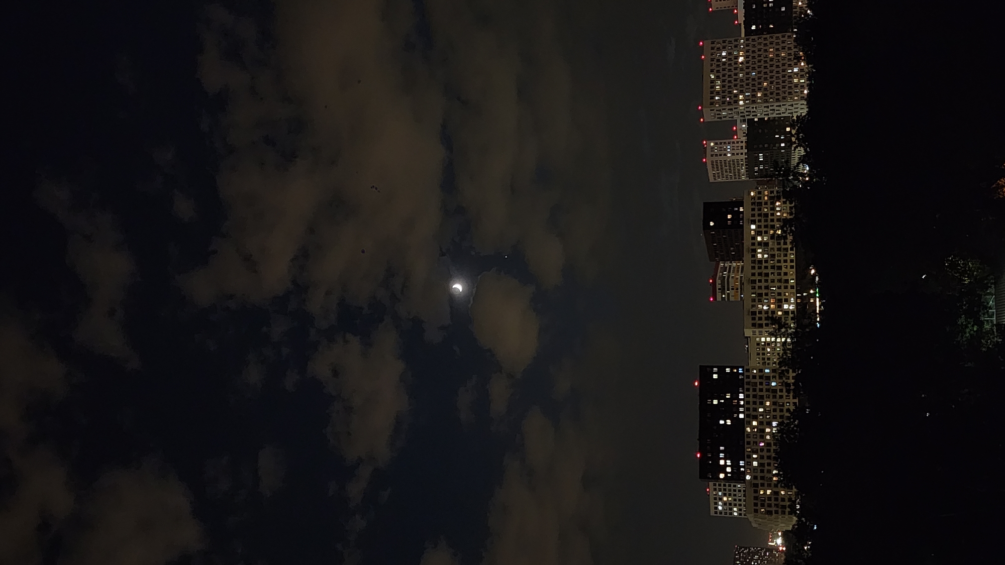 The image shows a nighttime scene in Moscow, Russia, with a crescent moon partially obscured by clouds. Some buildings are visible in the background. The sky is dark with yellow edges of the clouds. The buildings are light with yellow windows. Some trees are on the right and they're black.