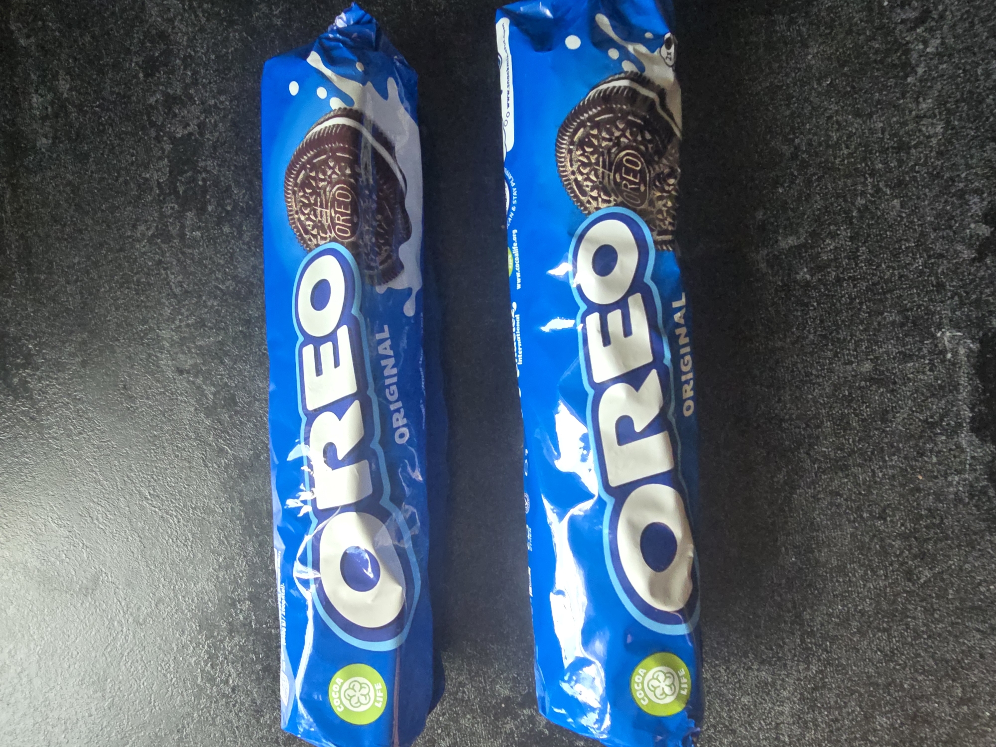 Two blue packages of Oreo cookies are lying side-by-side on a dark, textured surface. The packages are oriented vertically, with the top of the packages towards the top of the image. The front of each package prominently displays the "OREO" logo in large white letters with blue outlines. Each package also features an image of an Oreo cookie.
The package on the left is slightly angled towards the viewer. The package on the right is more directly facing the camera. Both packages have a graphic of milk splashing behind the Oreo cookie. At the bottom of each package, there is a green circular logo with the text "COCOA LIFE" and a clover-like symbol.
Visible text on the right package includes "ORIGINAL" below the Oreo logo, and some smaller text along the right edge which appears to be a website address, "www.cocoalife.org", and other unreadable text. The surface beneath the cookies appears to be a dark countertop or table with a speckled texture. There are no people or animals in the image. The lighting suggests an indoor setting, possibly in daylight, with the light source coming from above.