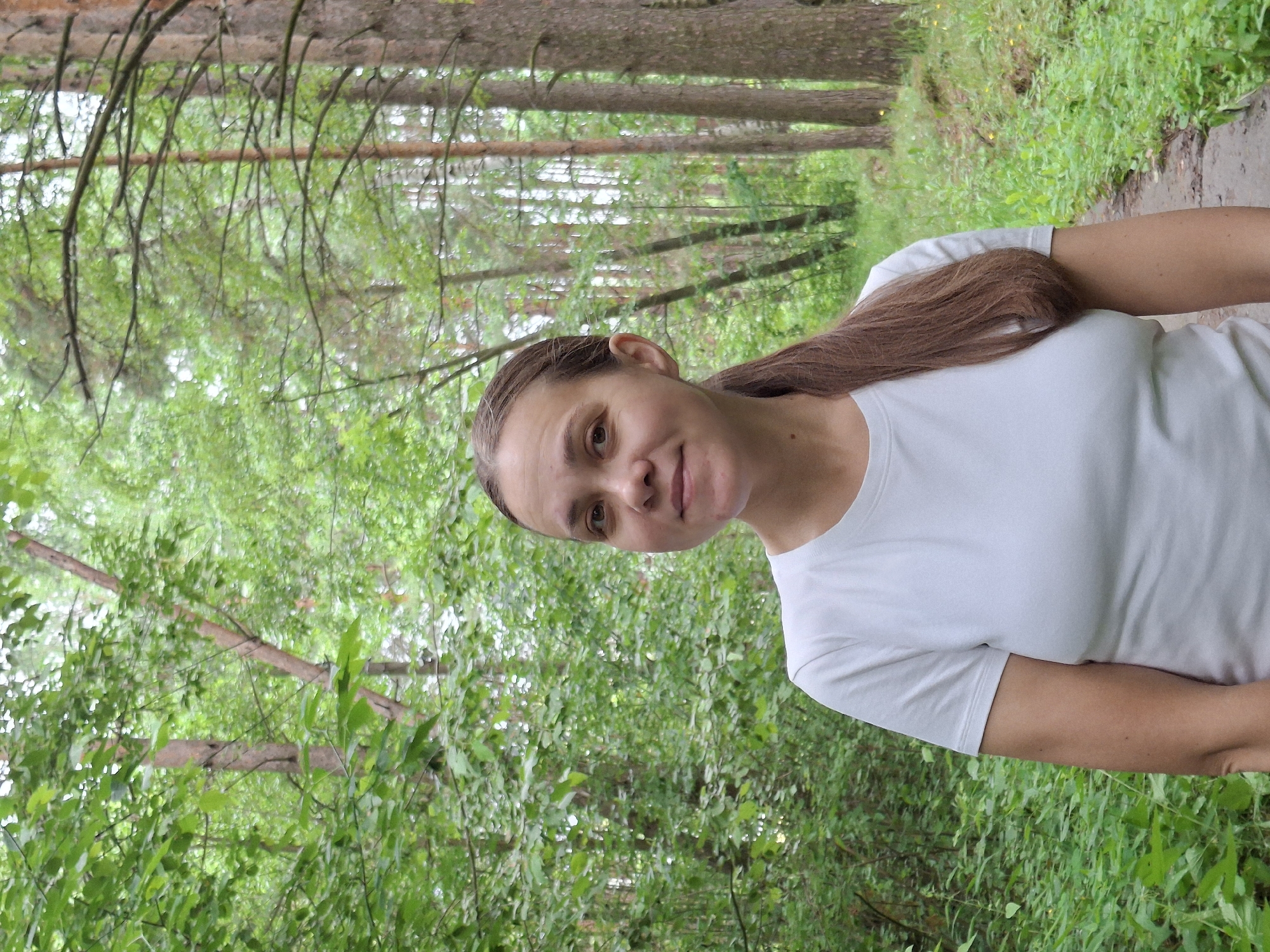 A woman with brown hair in a ponytail, wearing a white t-shirt, stands facing the camera in a forest. She is smiling slightly. The background is filled with lush green trees and foliage, suggesting a natural, outdoor setting. A dirt path is visible to her right. The image appears to be taken in the city of Chaikovsky, Russia, as per the user's request.